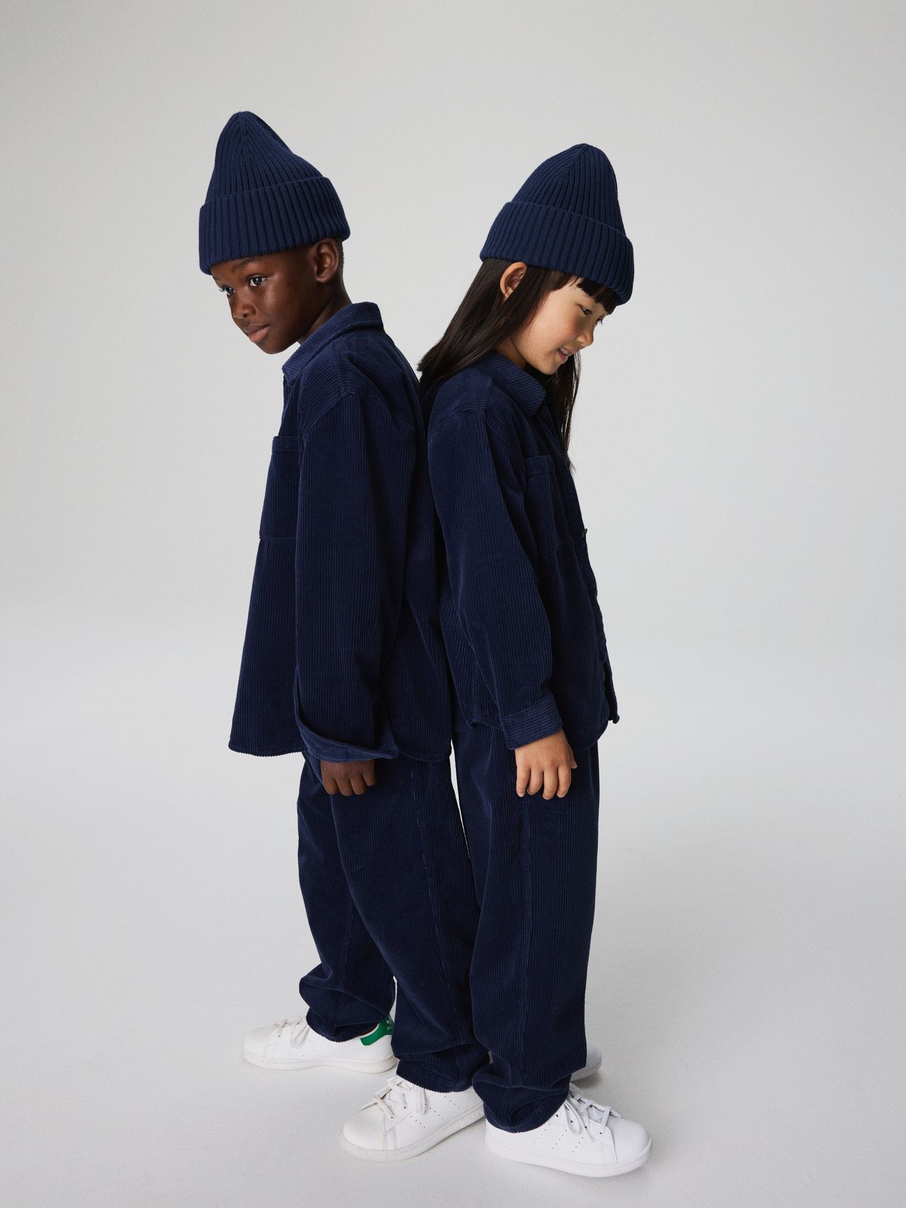 Two children stand back-to-back, wearing matching navy outfits and beanies, with white sneakers, against a plain background.