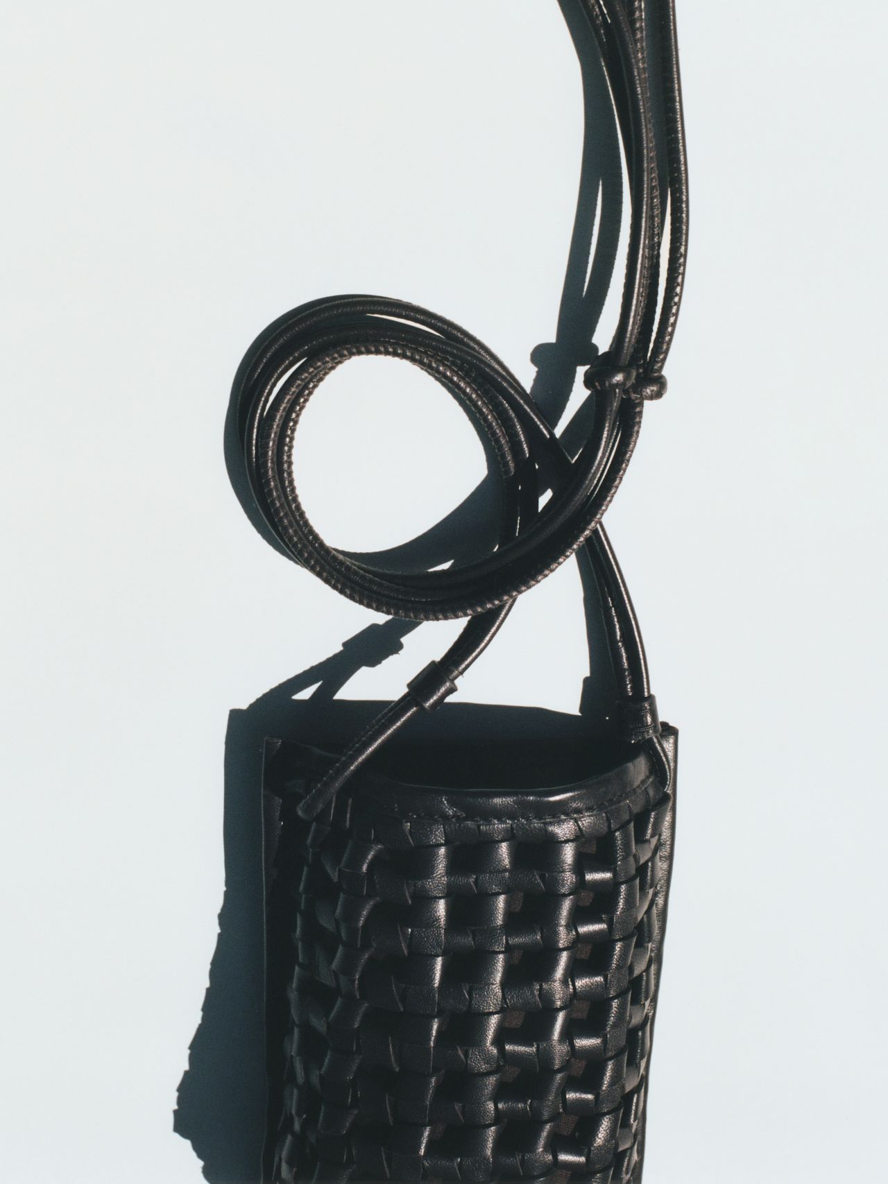 A black woven leather bag with a long, coiled strap against a light background, casting a shadow.