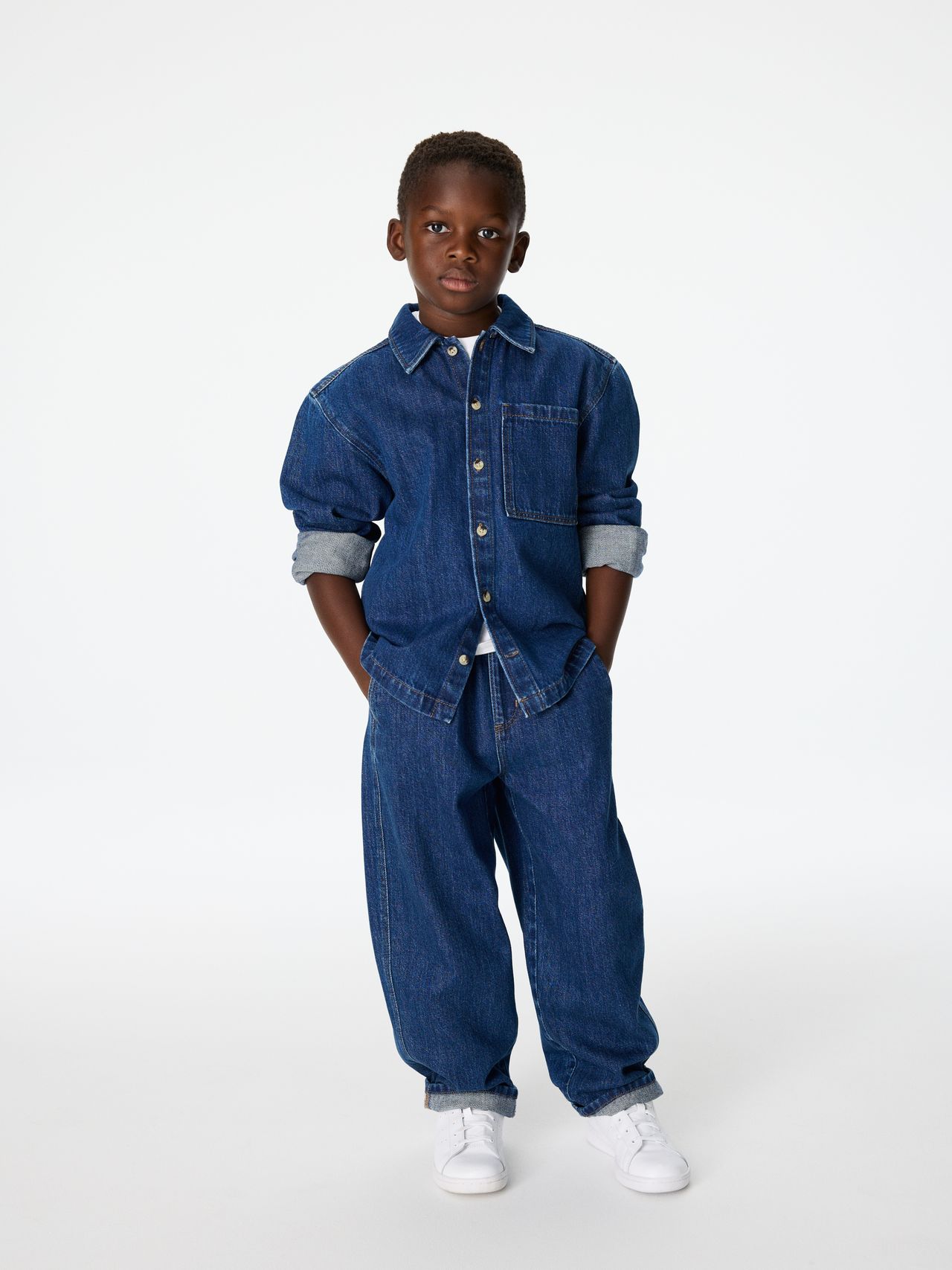 Young boy wearing a denim shirt and jeans, with rolled-up sleeves, standing confidently against a plain white background.