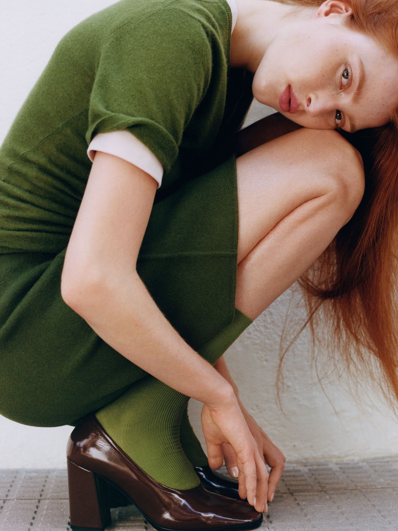 Woman in a green dress and matching socks squats, wearing brown heels, against a light background.