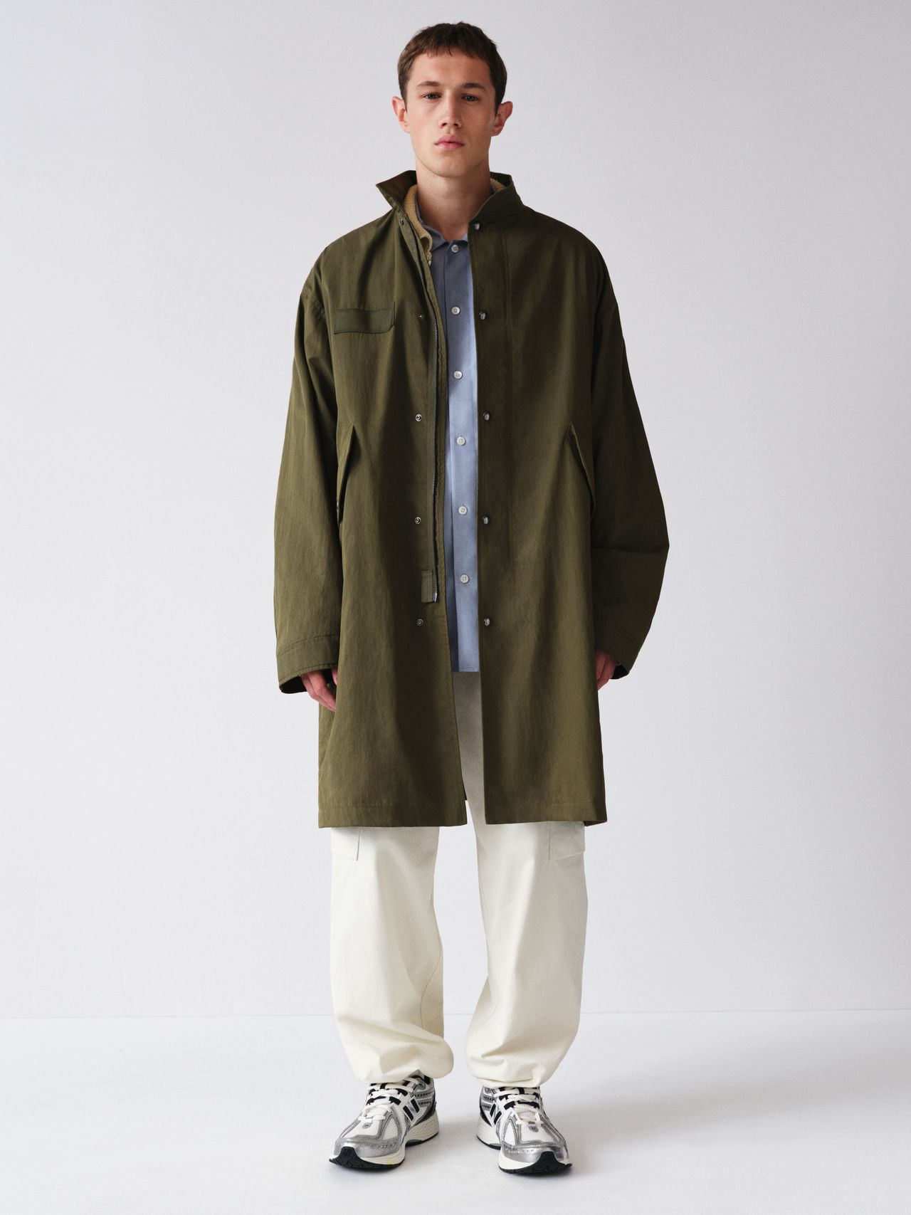 Person wearing a green coat over a blue shirt, white pants, and sneakers, standing against a plain white background.