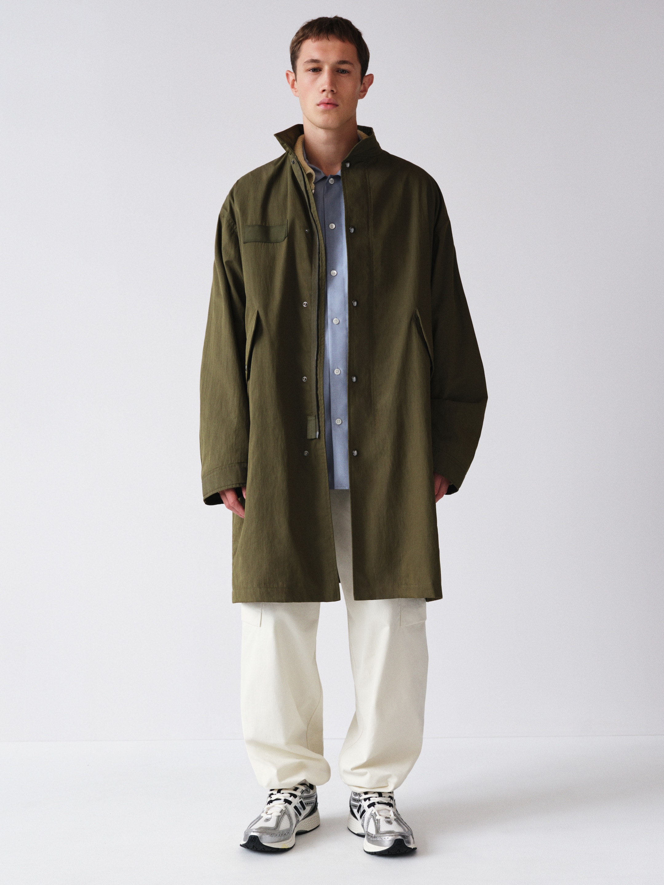 Person wearing a green coat over a blue shirt, white pants, and sneakers, standing against a plain white background.