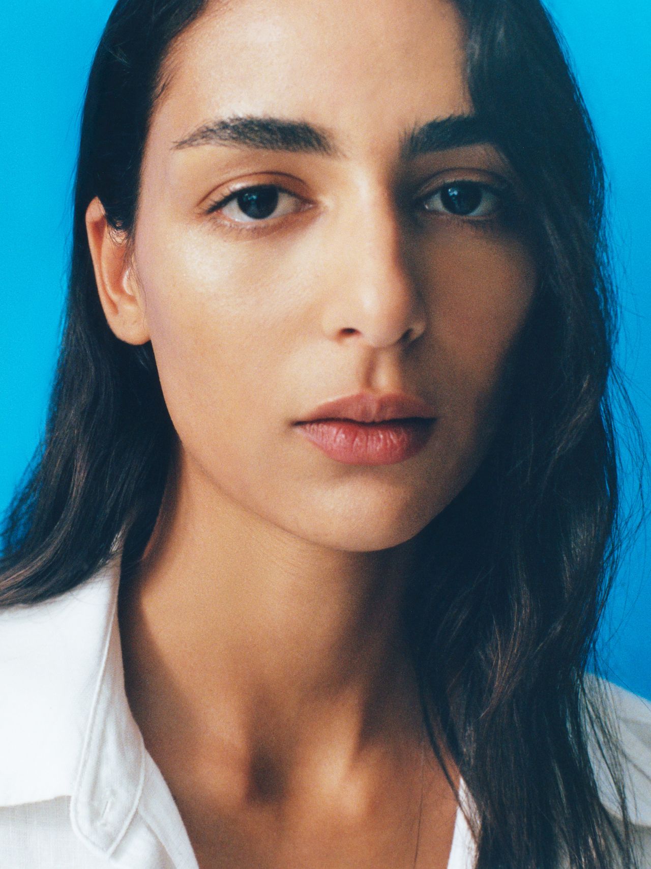 A person with long dark hair and a white shirt looks directly at the camera against a bright blue background.