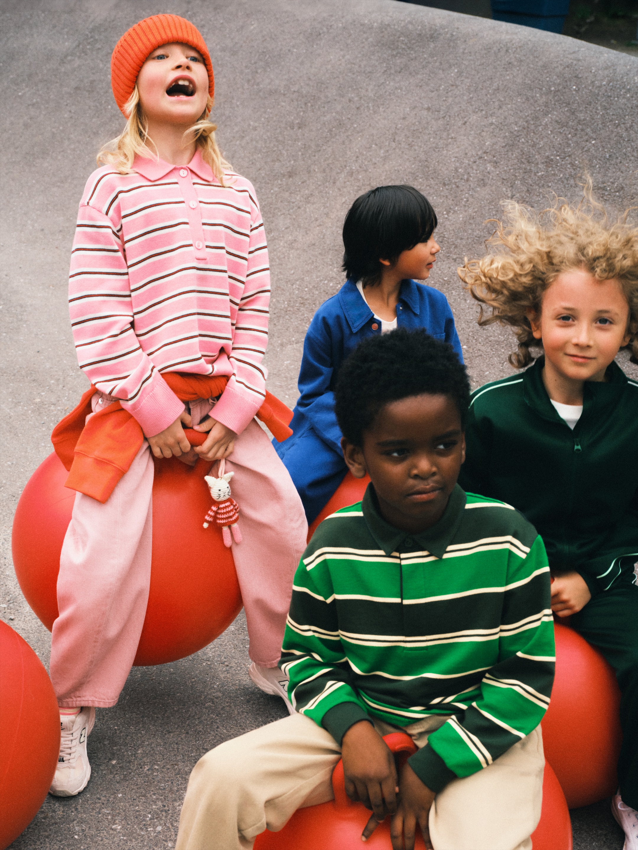 Four children sitting on red bouncing balls, wearing colorful clothing and smiling, with one child looking off to the side.
