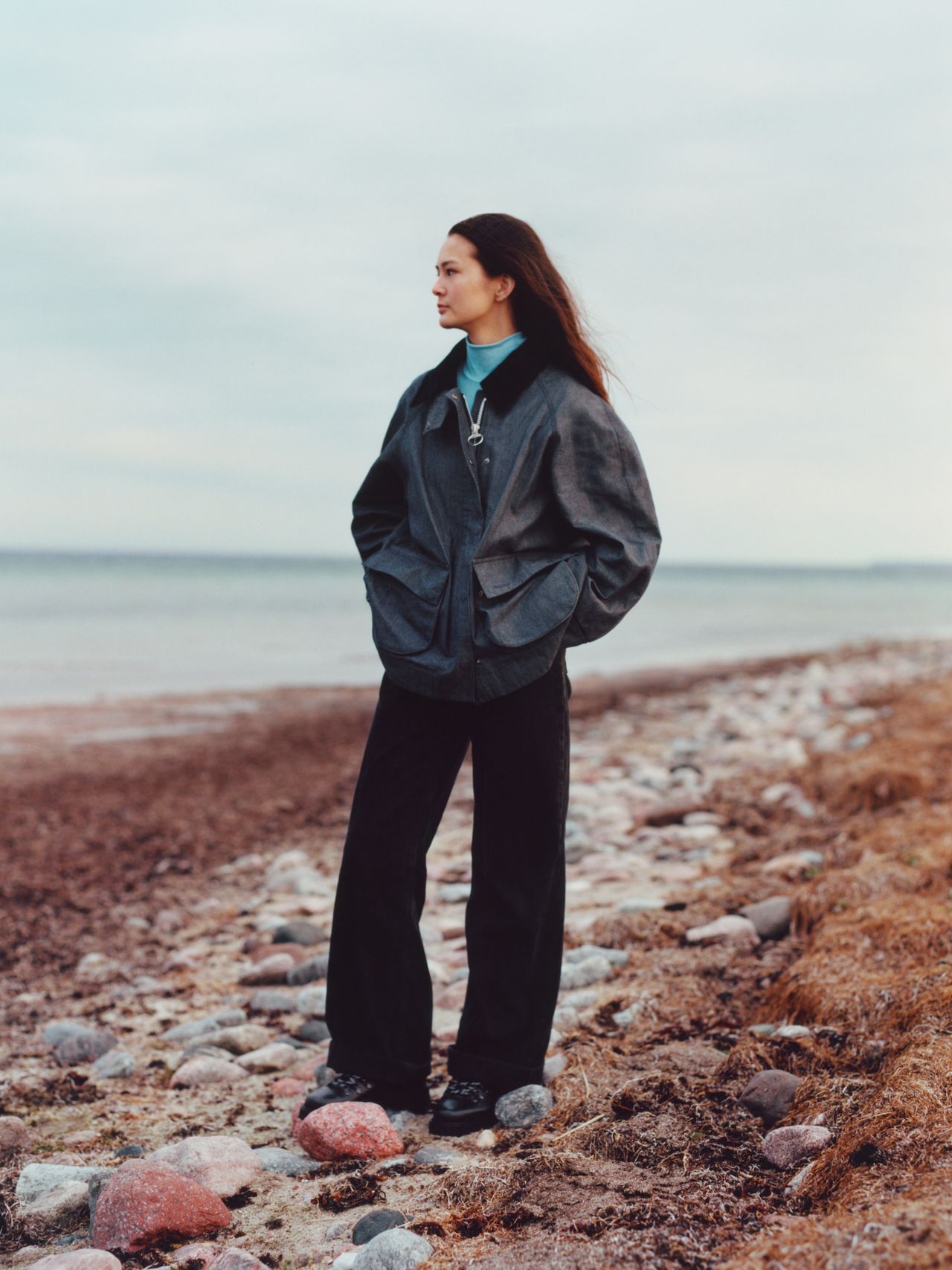 A person stands on a rocky beach wearing a dark jacket and pants, looking into the distance under a cloudy sky.