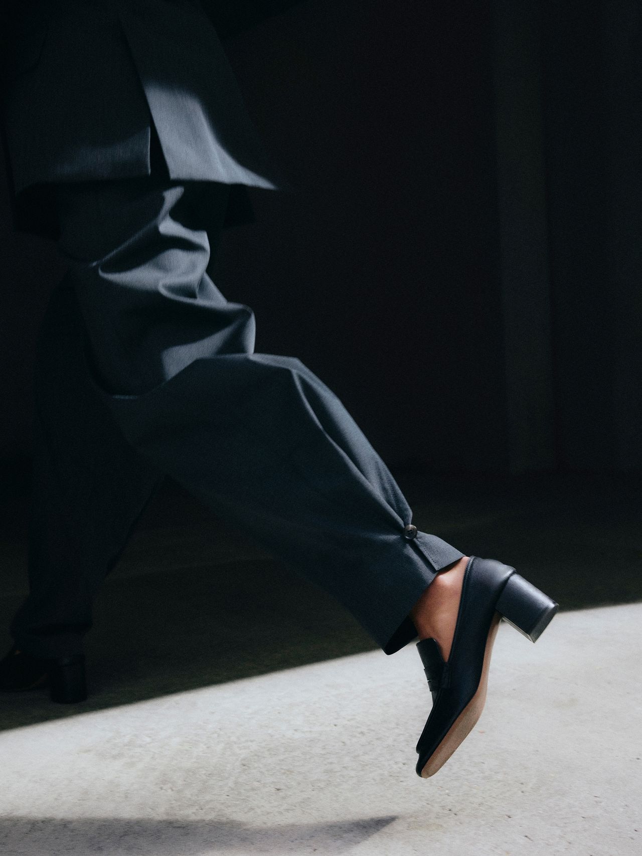 Person in dark suit walking, showing only legs and feet, wearing black heeled shoes, with sunlight casting shadows on the ground.