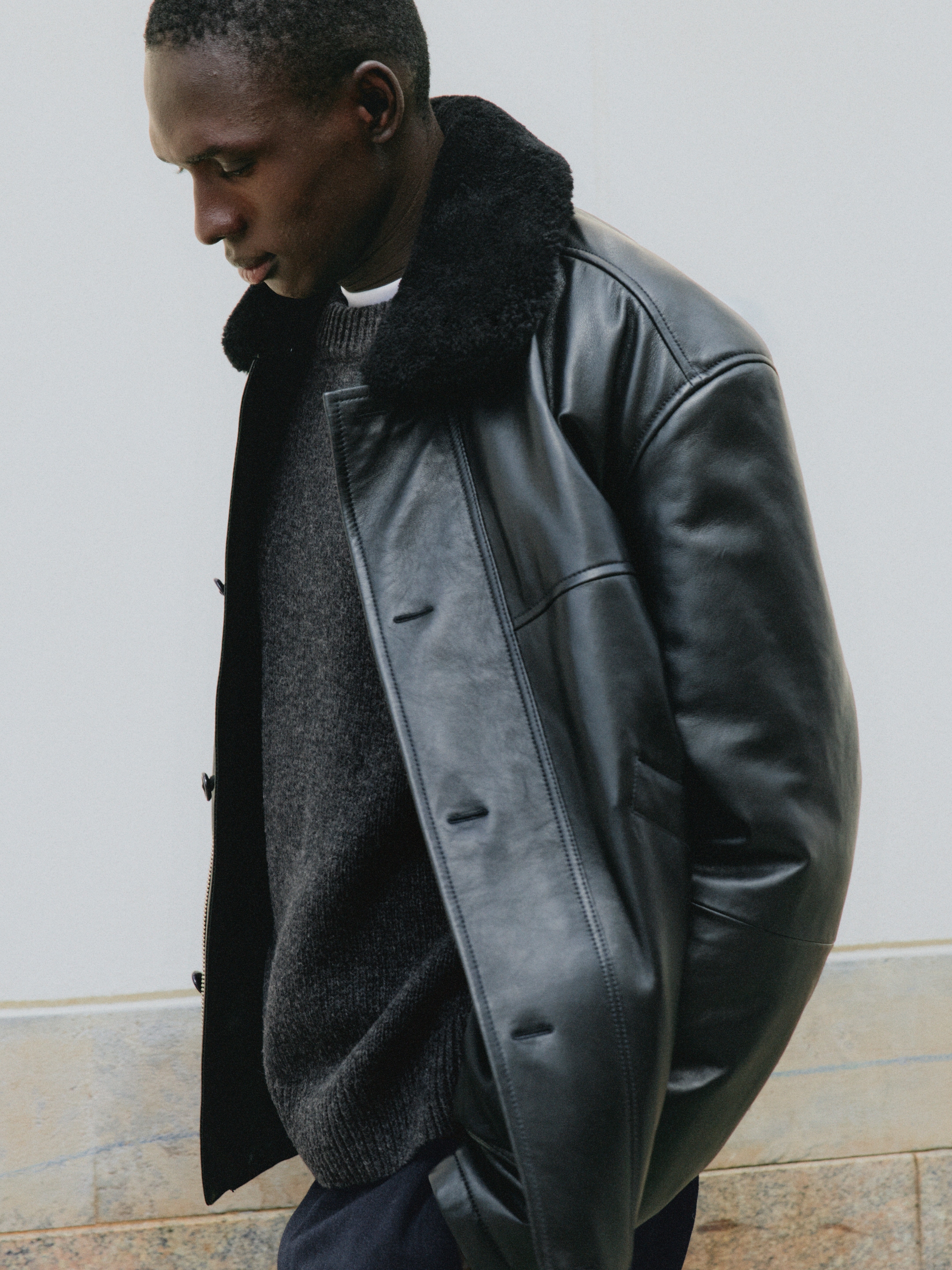 Man in a black leather jacket with a fur collar looking down, wearing a dark sweater against a light background.