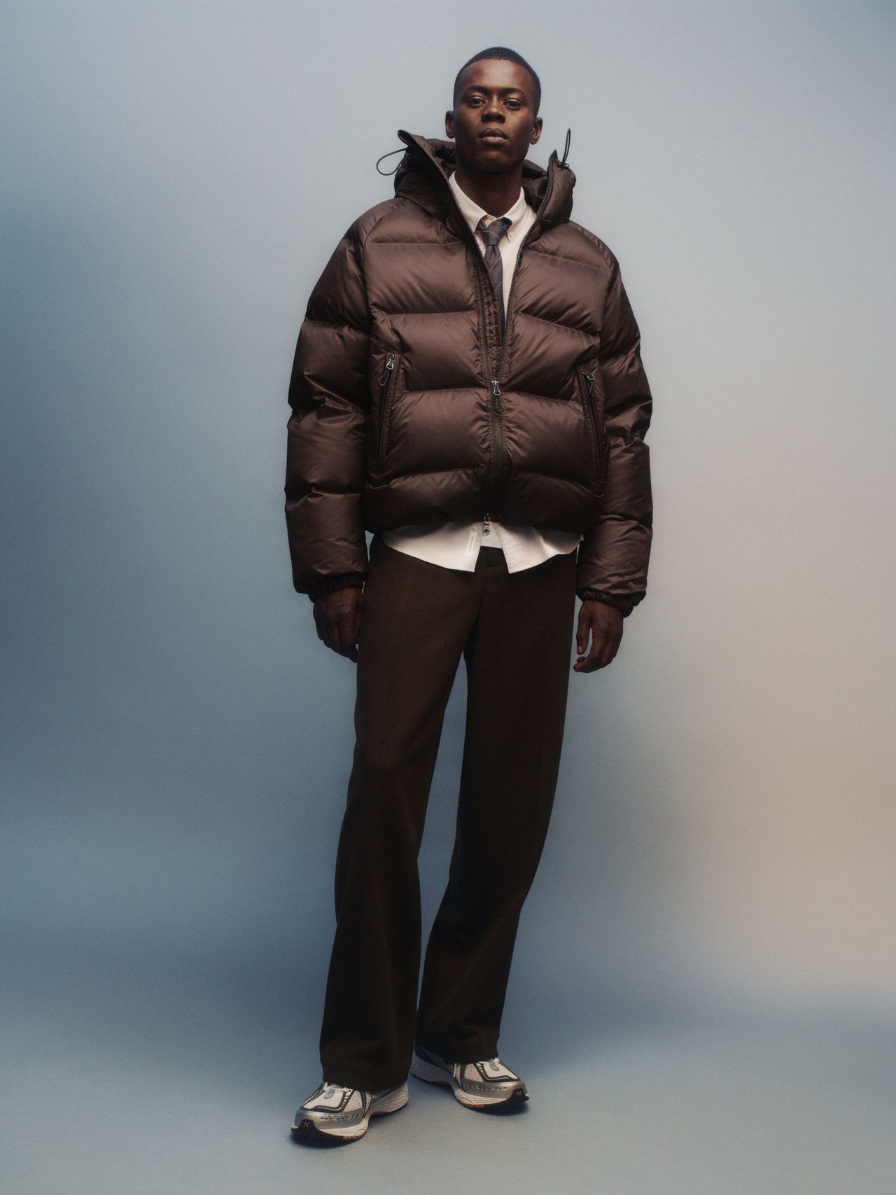 Person wearing a dark puffer jacket, white shirt, tie, dark pants, and sneakers, standing against a gradient blue background.