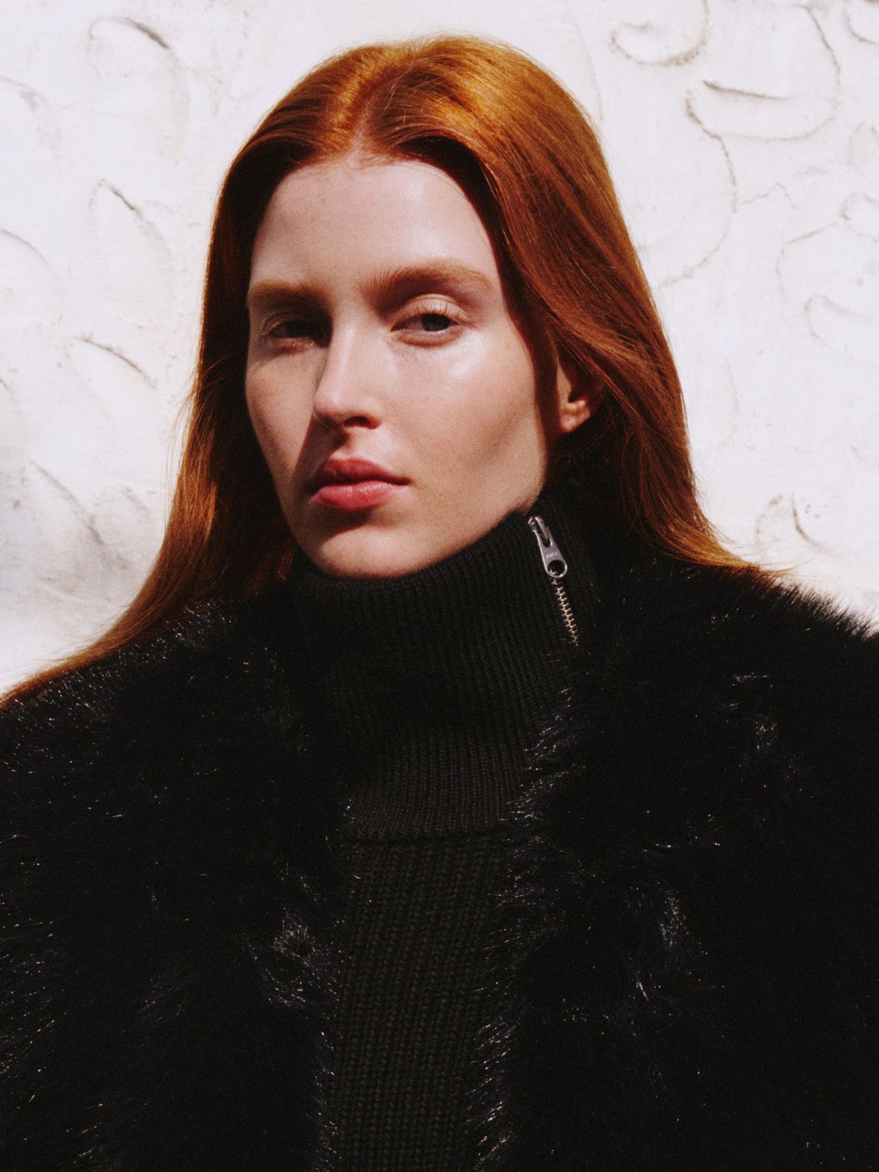 Person with long red hair wearing a black fur coat and turtleneck, standing against a textured white wall.