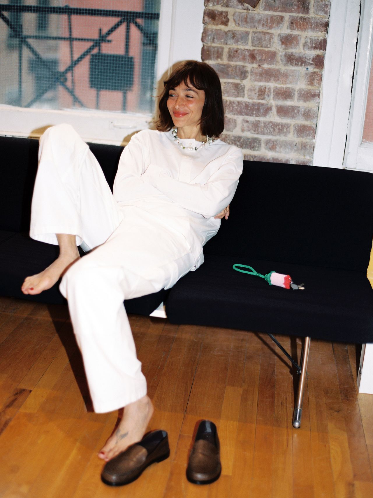A person in a white outfit sits relaxed on a black sofa, barefoot, with brown shoes on the floor and a colorful keychain nearby.
