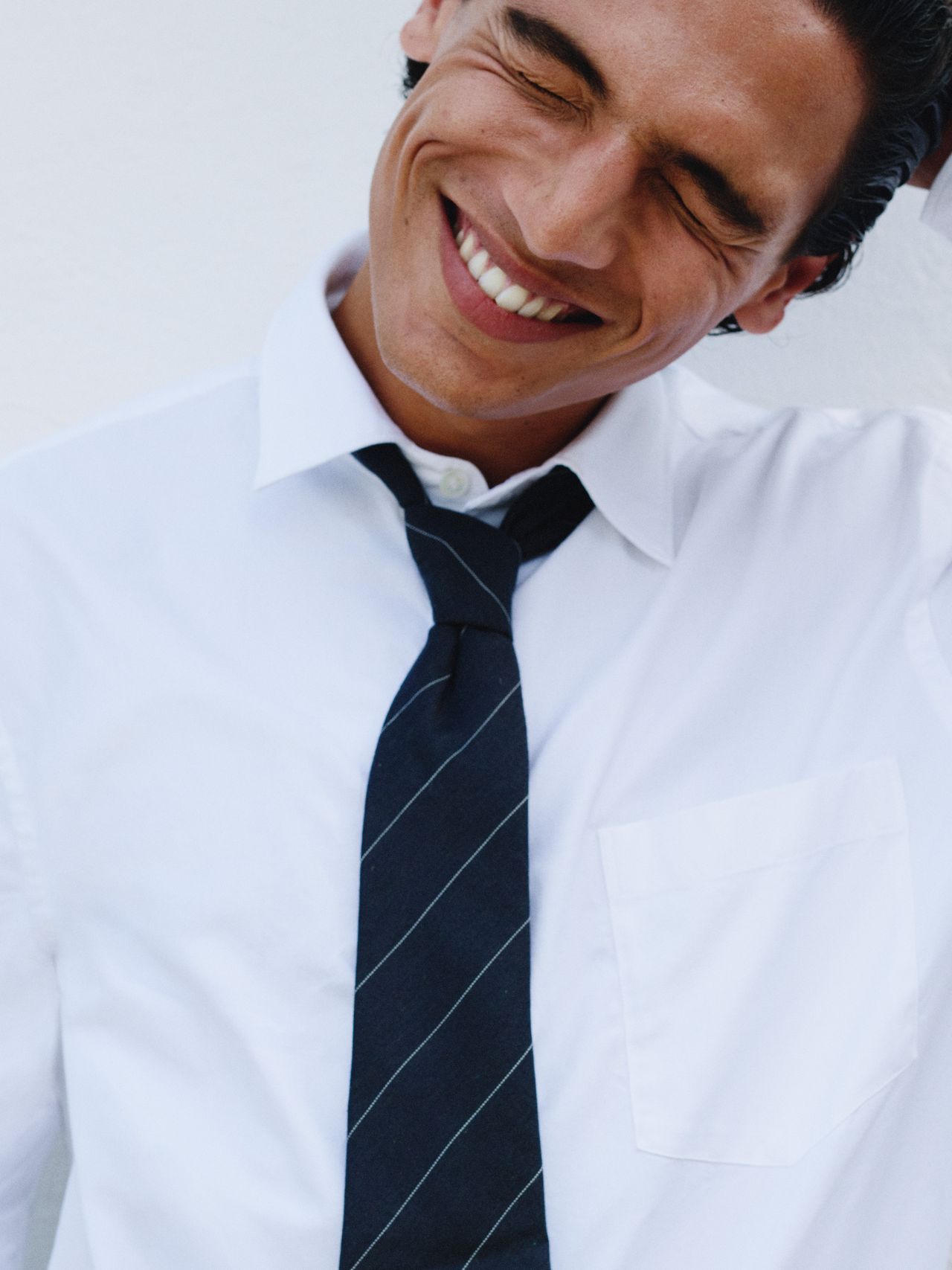 Smiling man in a white shirt and striped tie, eyes closed, standing against a white background.