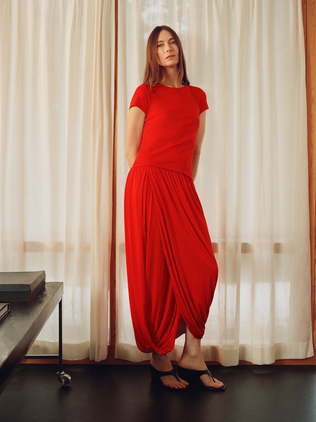 A person stands in front of beige curtains wearing a red outfit with draped pants and black shoes, hands behind their back.