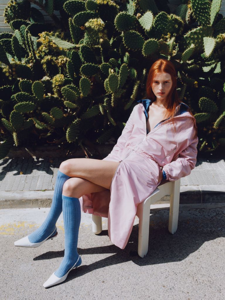A person with long red hair sits on a white chair, wearing a pink outfit, blue knee-high socks, and white shoes, with cacti in the background.