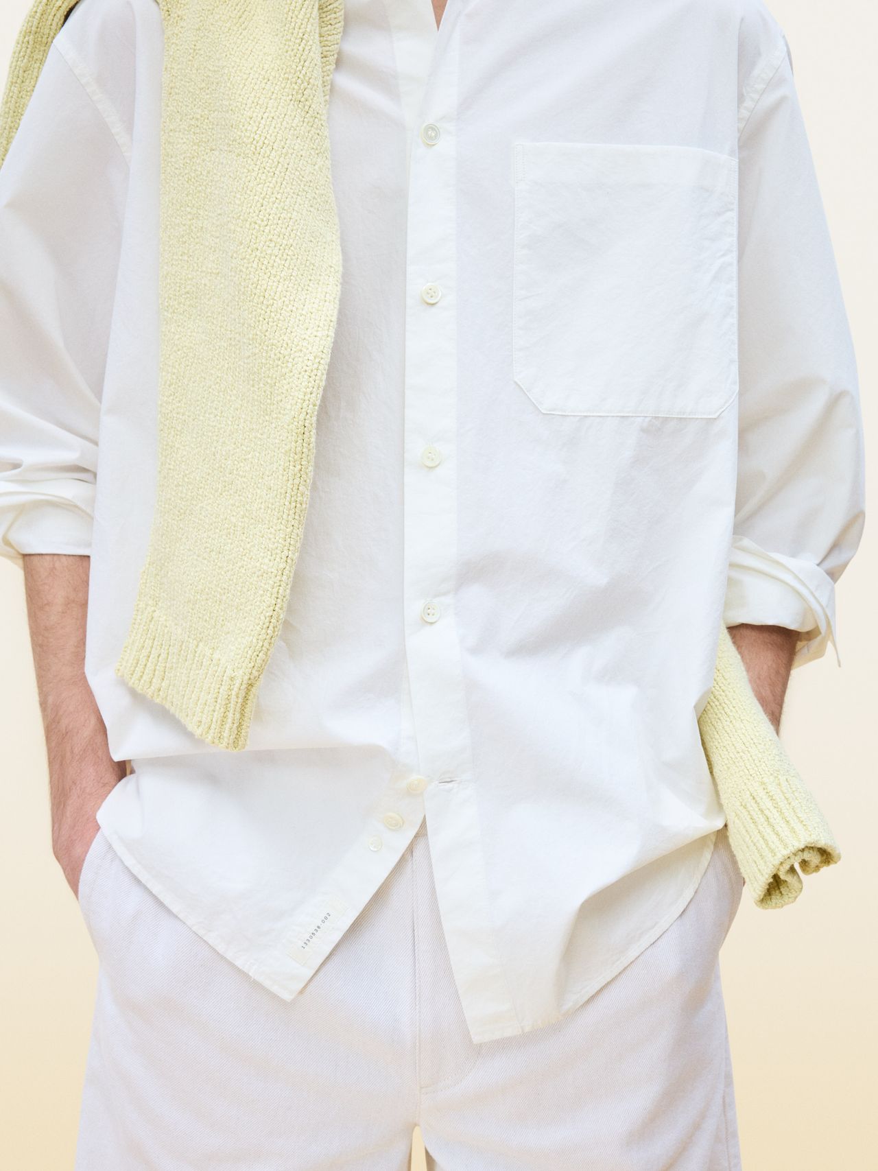 Person wearing a white button-up shirt with a yellow sweater draped over shoulders, hands in pockets, against a neutral background.