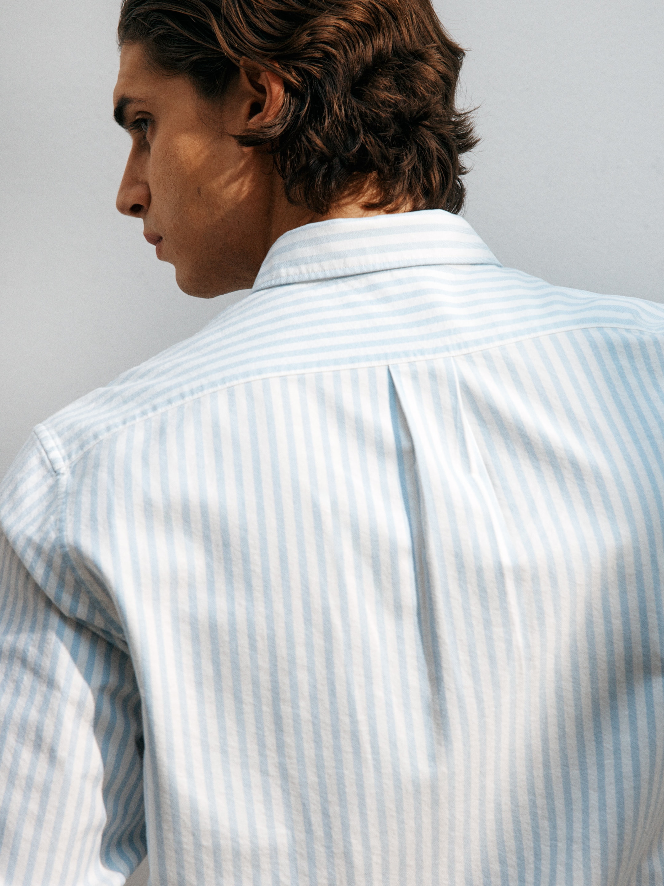 Person with wavy hair in profile, wearing a light blue and white striped shirt, looking contemplative against a plain background.