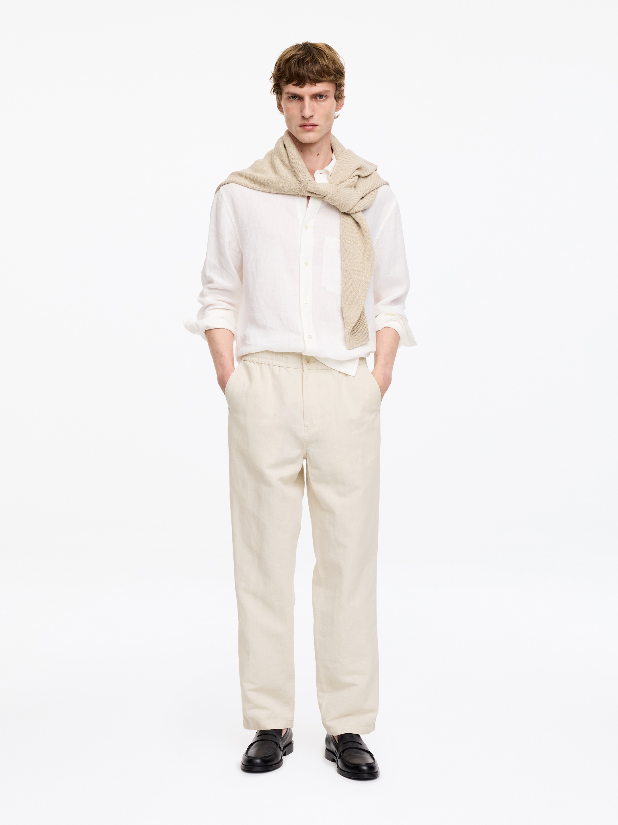 Young man in a white shirt and beige pants with a matching sweater draped over shoulders, hands in pockets, standing against a white background.