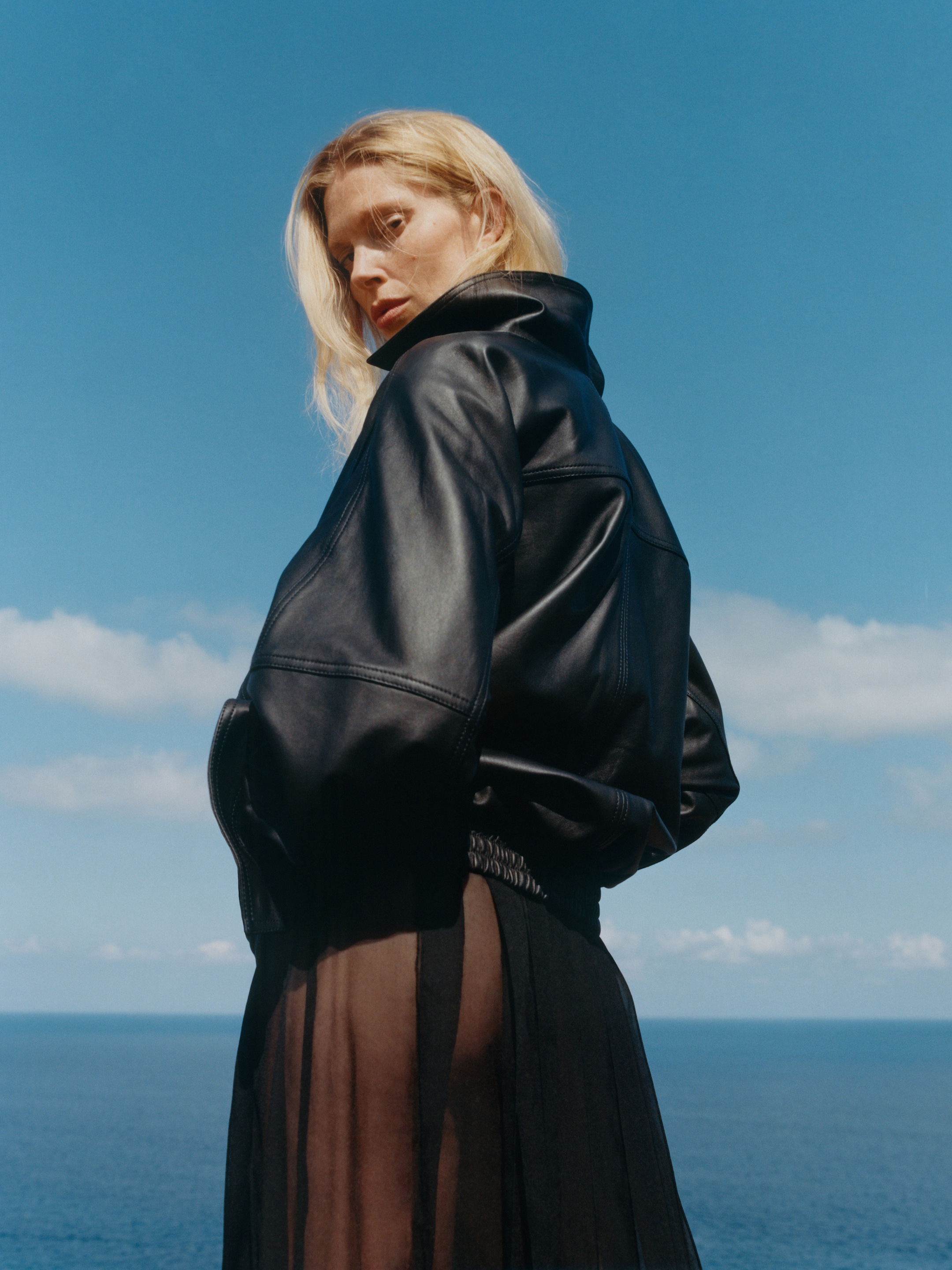 Woman wearing a black leather jacket and sheer skirt stands by the ocean, looking back over her shoulder against a clear blue sky.