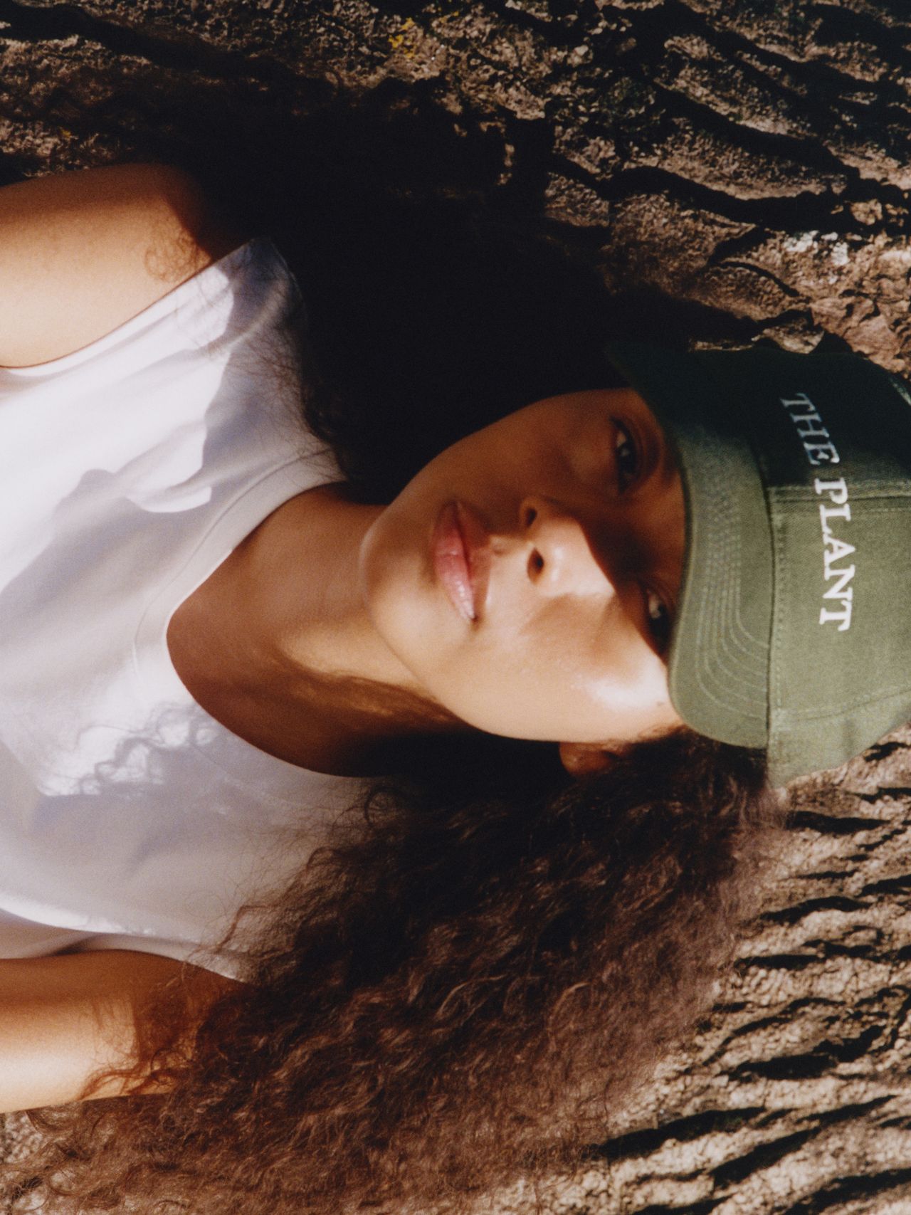Person with curly hair wearing a green cap and white shirt, lying on textured ground, looking up. Cap reads "THE PLANT."