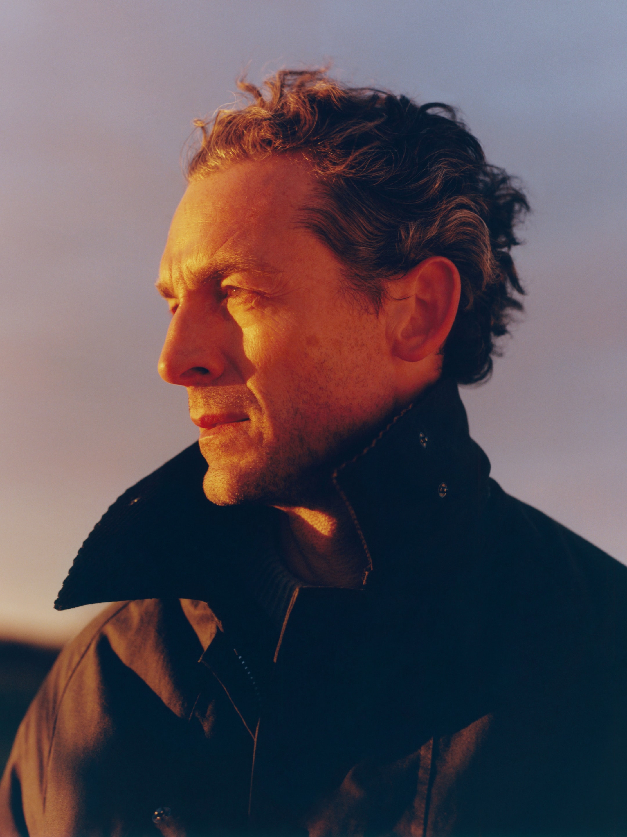 Man with curly hair in a dark coat gazes into the distance, illuminated by warm, golden light against a soft, blurred background.