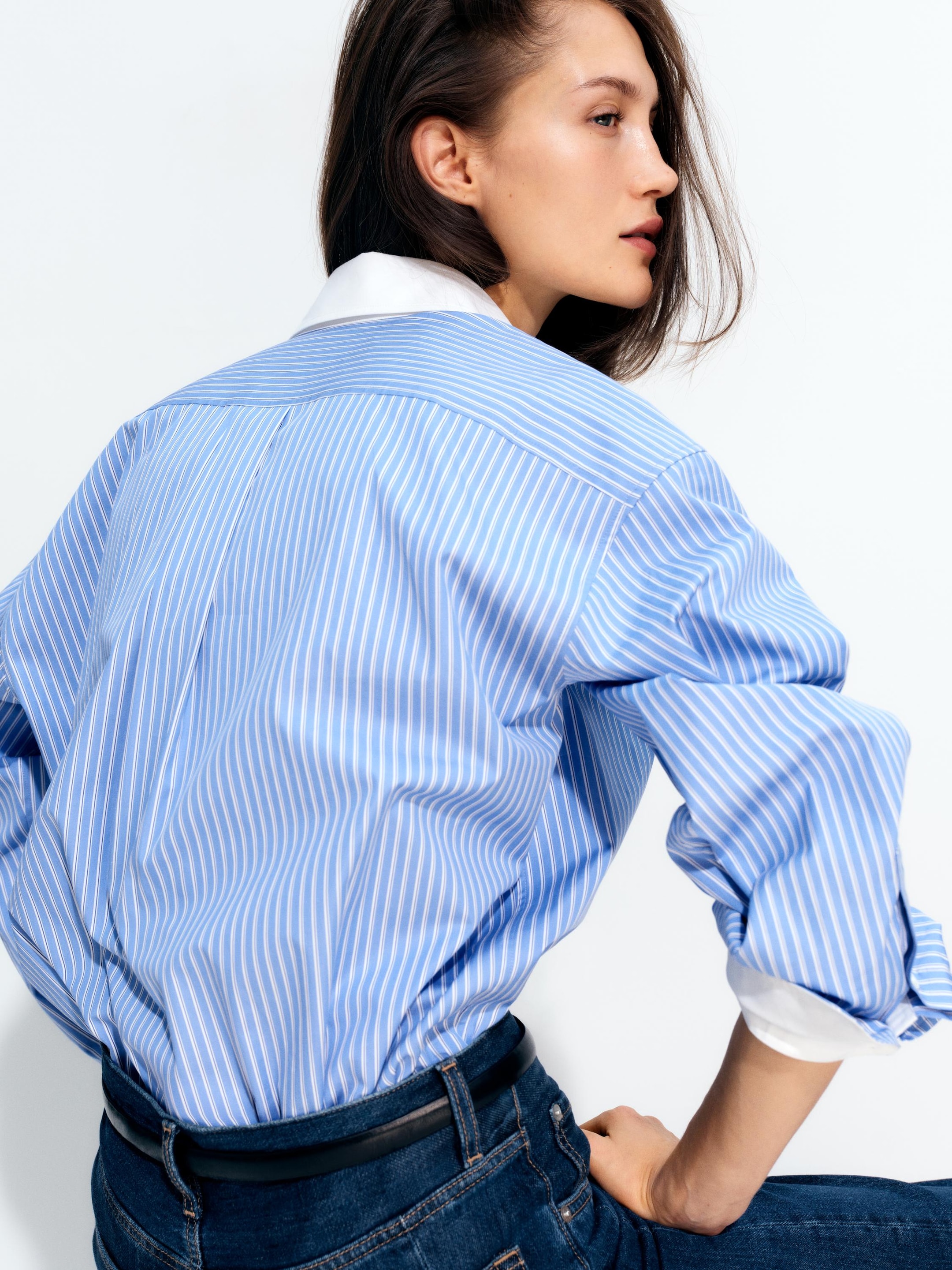 A person wearing a blue and white striped shirt, with sleeves rolled up, sits sideways against a white background.