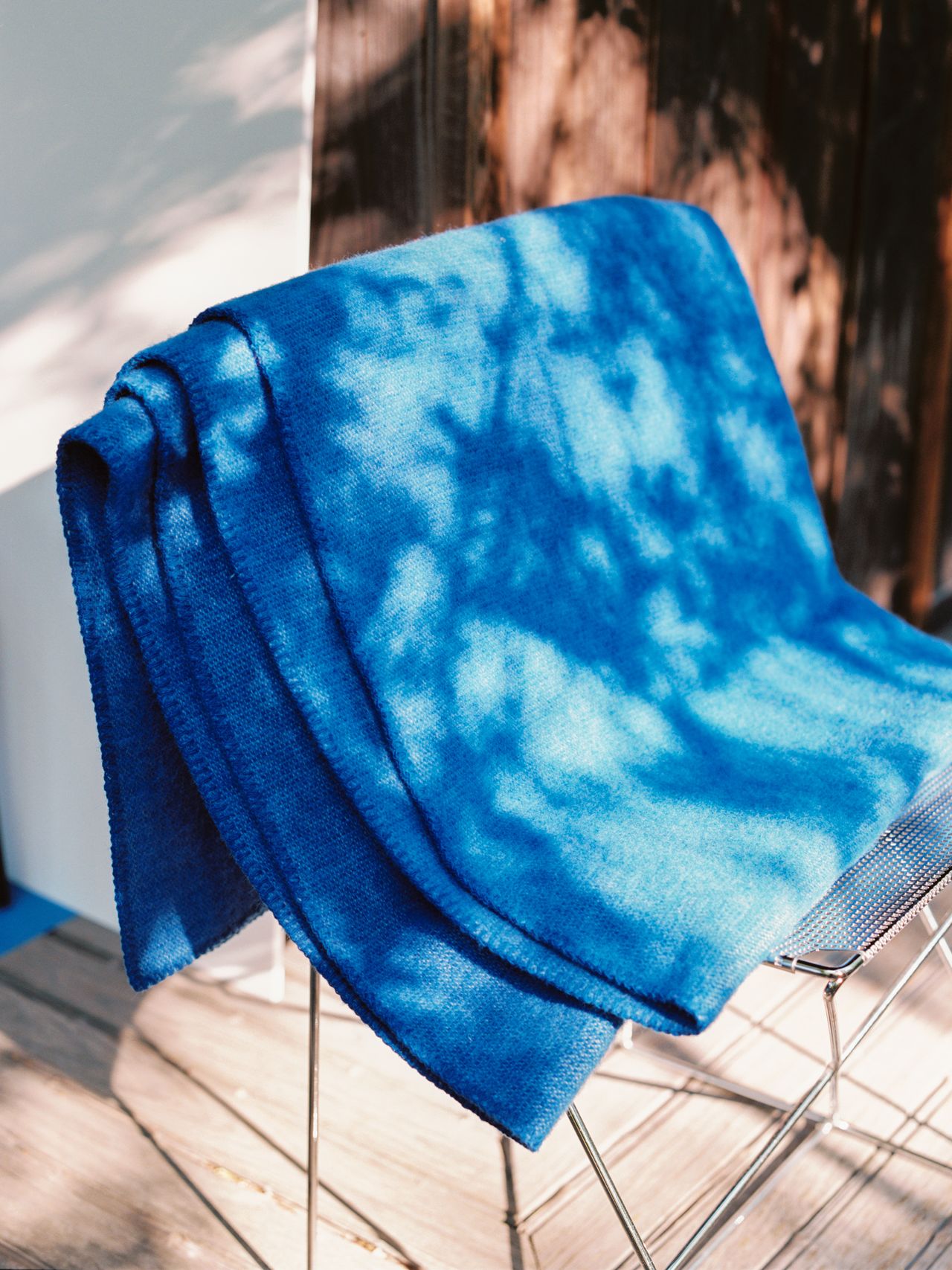 A blue blanket draped over a metal chair, with sunlight casting leafy shadows on it, set on a wooden deck.