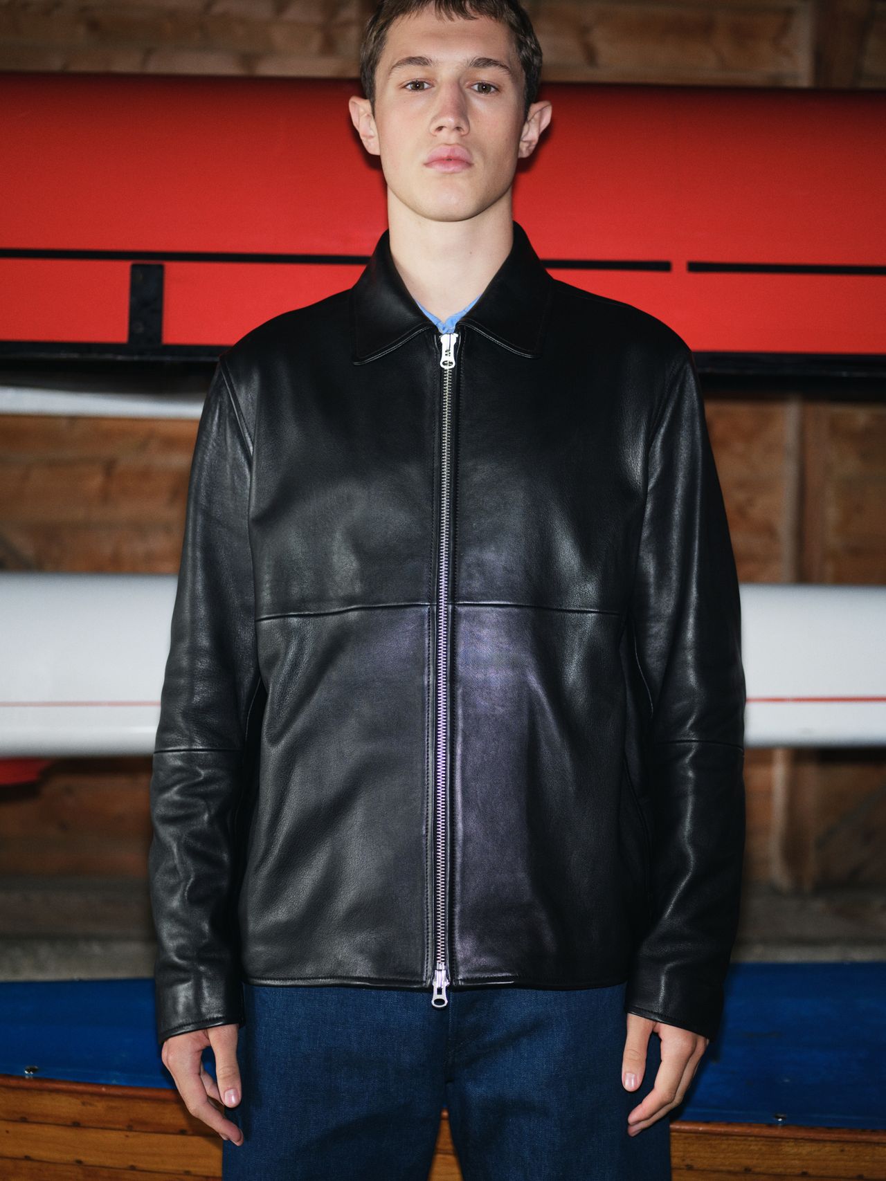 Person wearing a black leather jacket and blue jeans, standing indoors with red and white background elements.