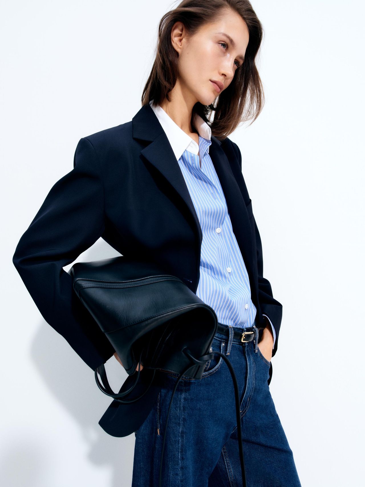 A person in a navy blazer and striped shirt holds a black bag, posing against a plain white background.