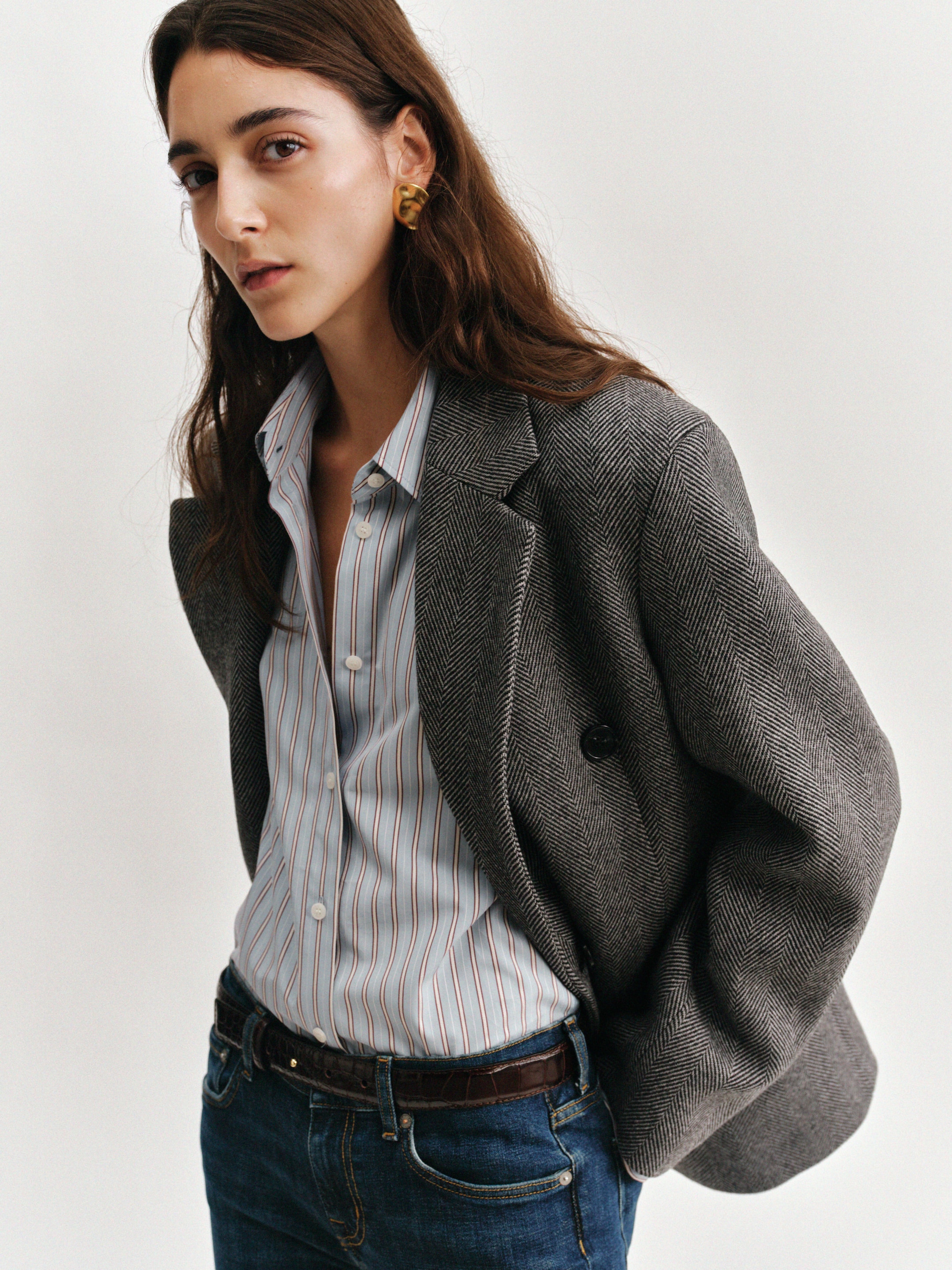 A person with long brown hair wearing a gray blazer, striped shirt, and jeans, looking confidently at the camera against a plain background.