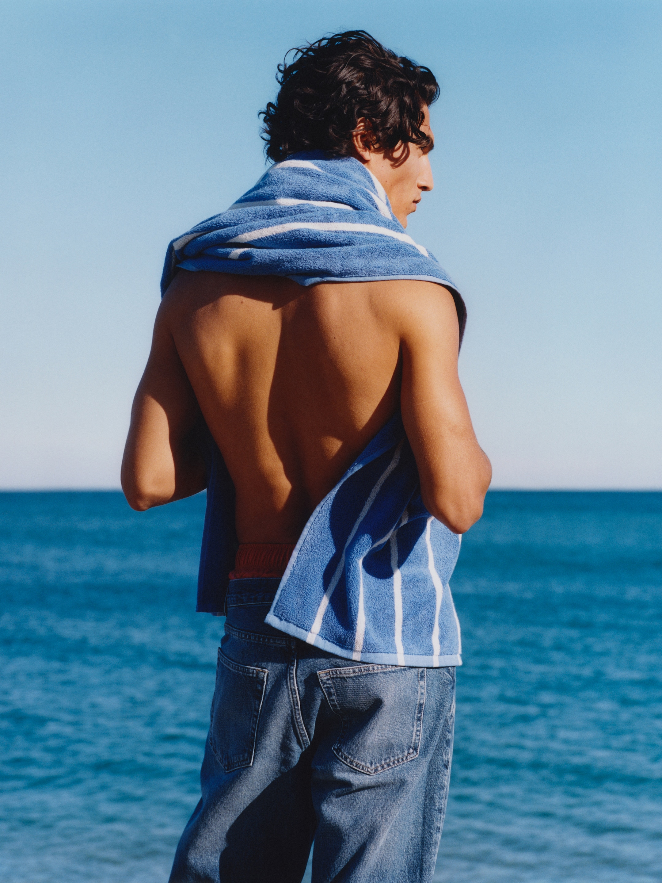 A person stands on a beach, facing the ocean, with a blue and white striped towel draped over their shoulders, wearing jeans.