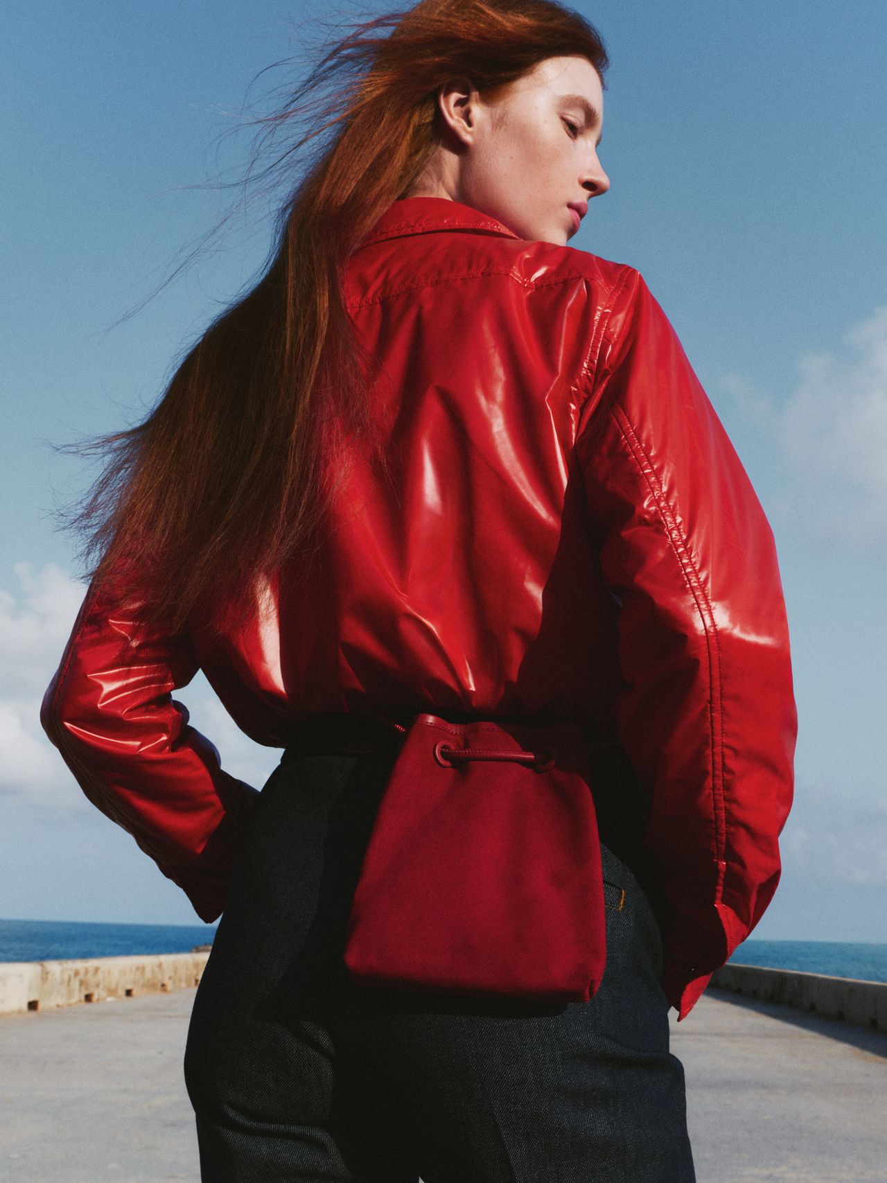 Woman with long red hair in a shiny red jacket and black pants stands on a pier, facing away, with a red bag against a blue sky.