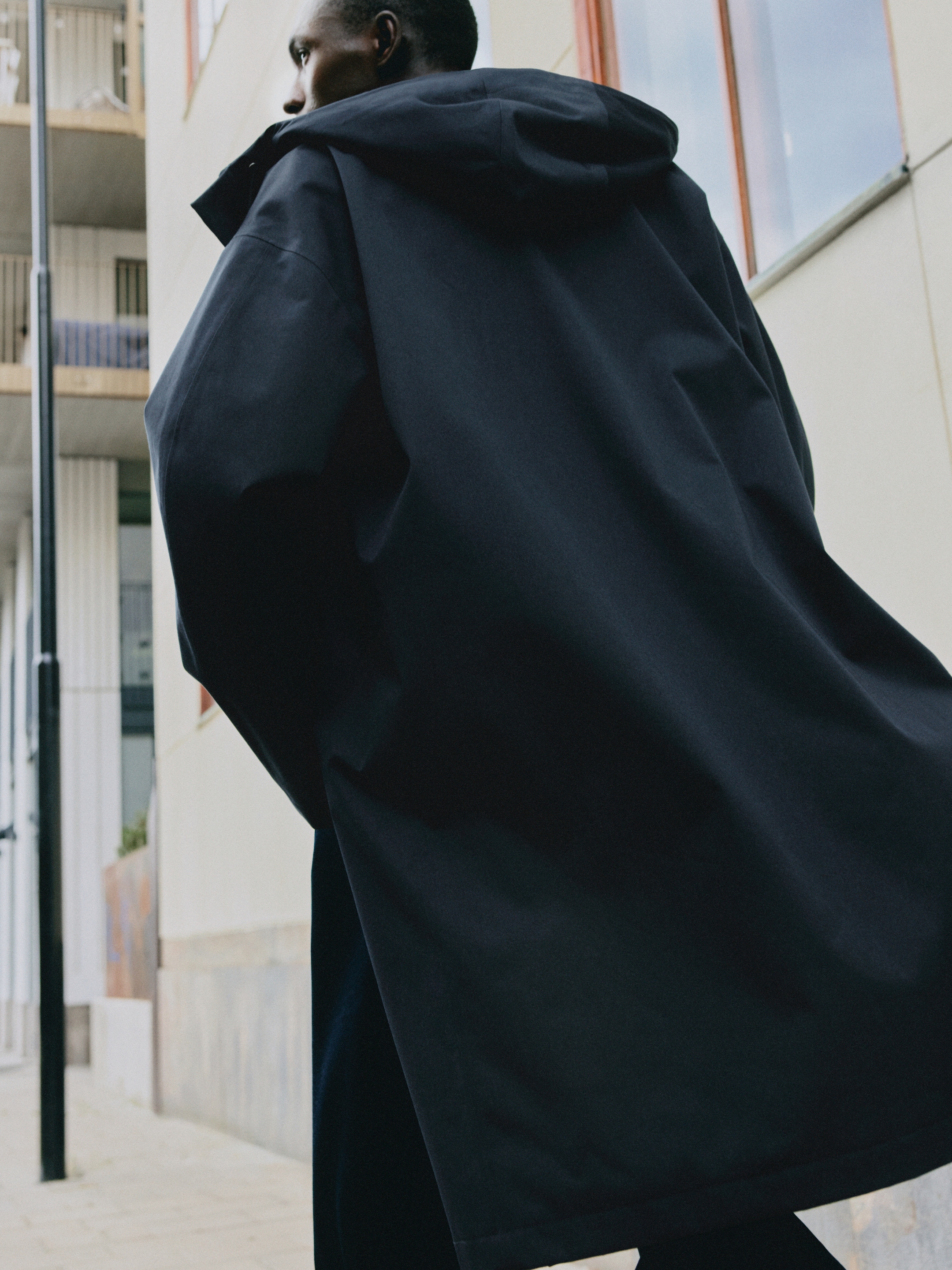 A person in a dark, oversized coat walking on a city sidewalk. Modern building with large windows in the background.