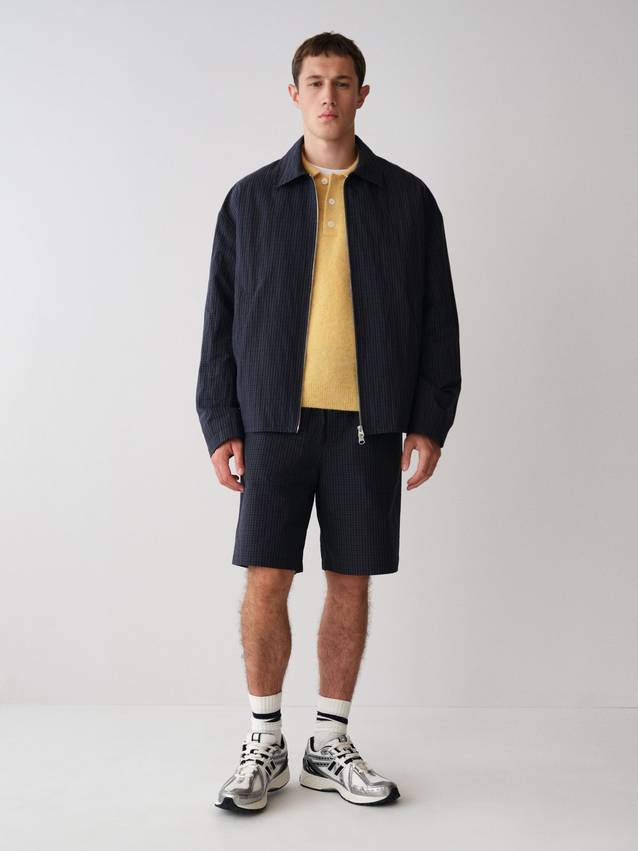 Man wearing a dark jacket, yellow shirt, and matching shorts, with white socks and sneakers, standing against a plain white background.