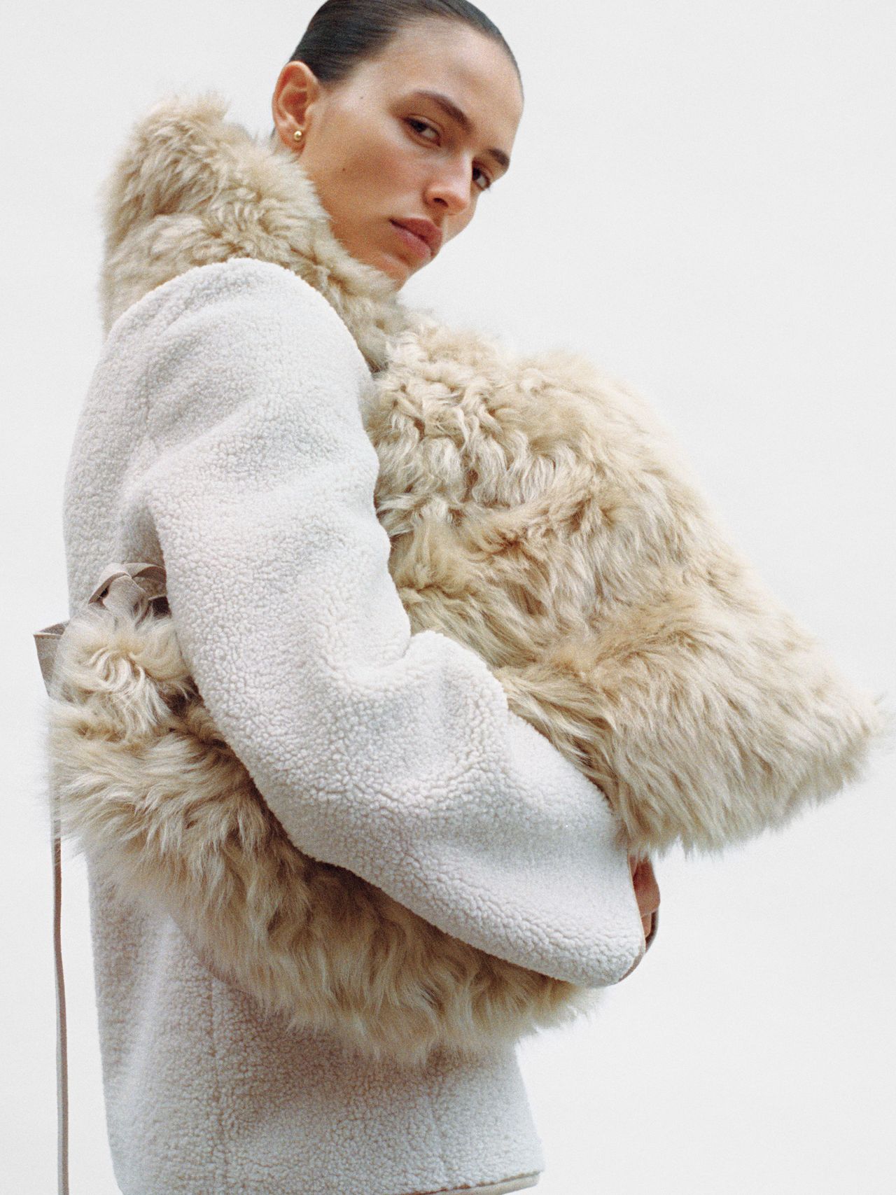 Person in a white textured coat holding a large, fluffy beige bag, looking over their shoulder against a plain background.