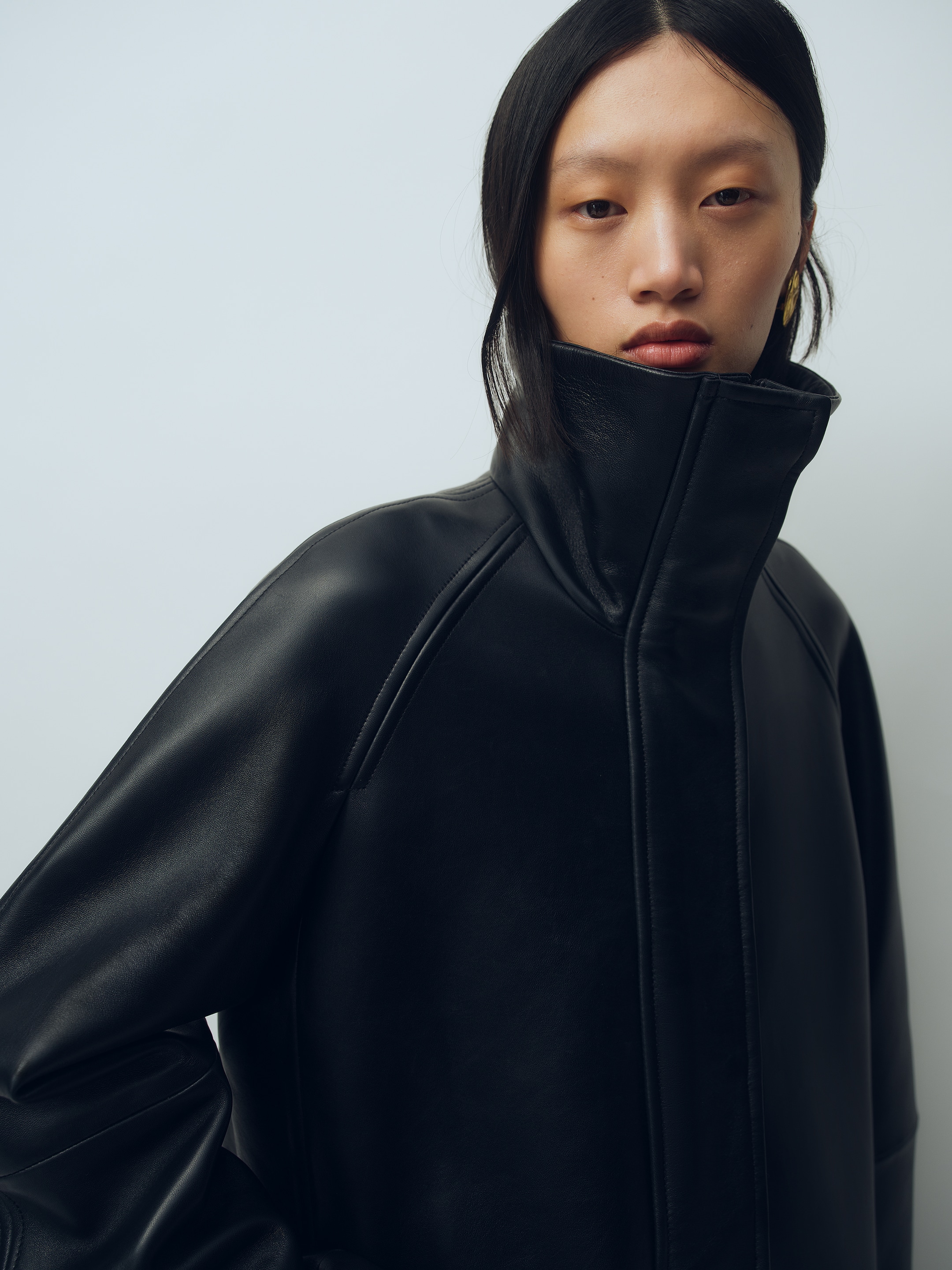 Person wearing a high-collared black leather jacket, standing against a light background, with serious expression and sleek black hair.