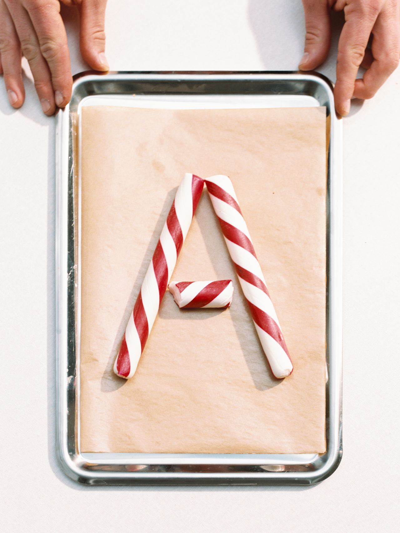 Red and white striped candy canes arranged in the shape of the letter "A" on a baking tray lined with parchment paper, with hands holding the tray.
