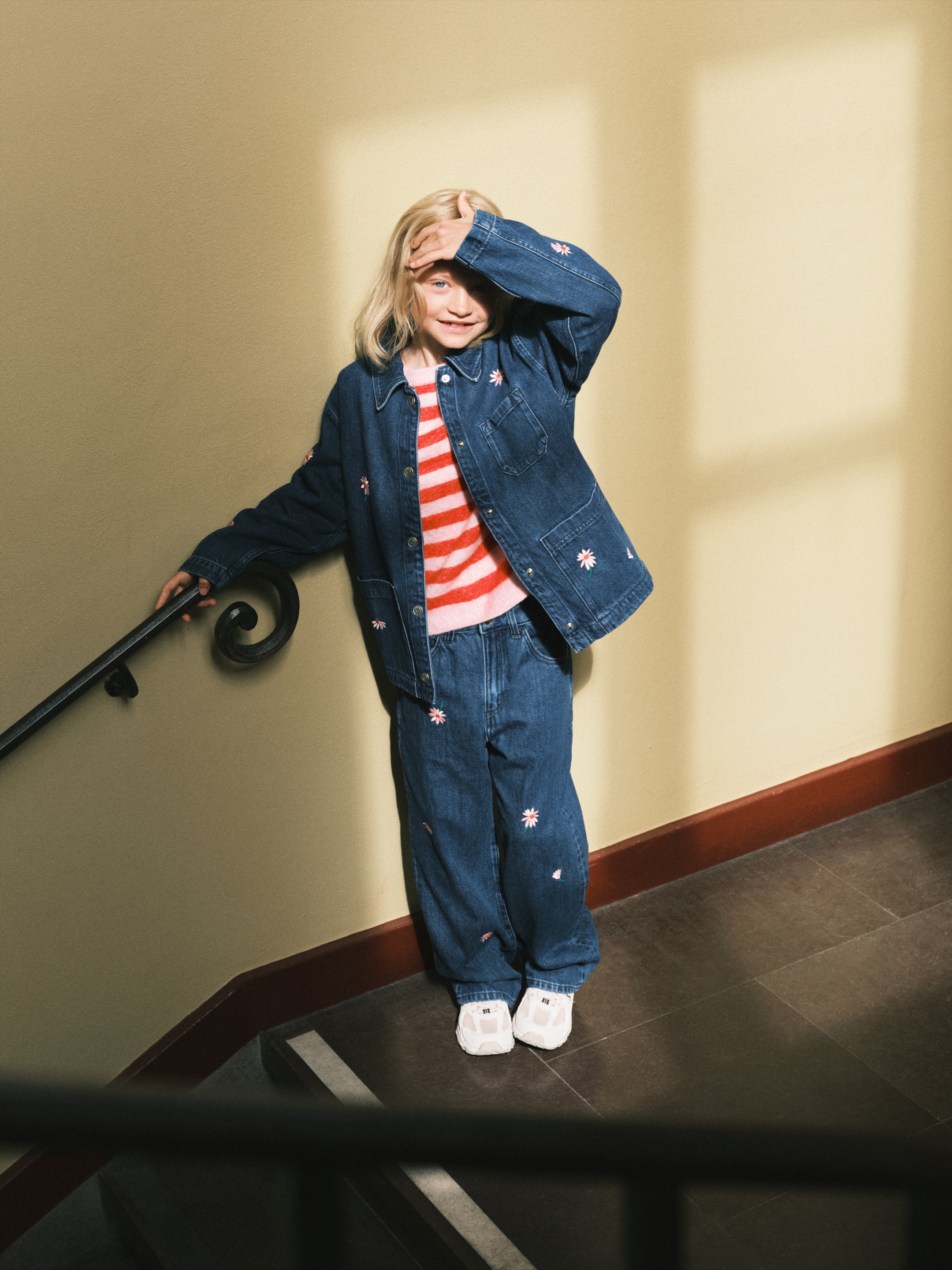Child wearing a denim jacket and jeans with floral embroidery, standing on a staircase and smiling, sunlight casting shadows on the wall.
