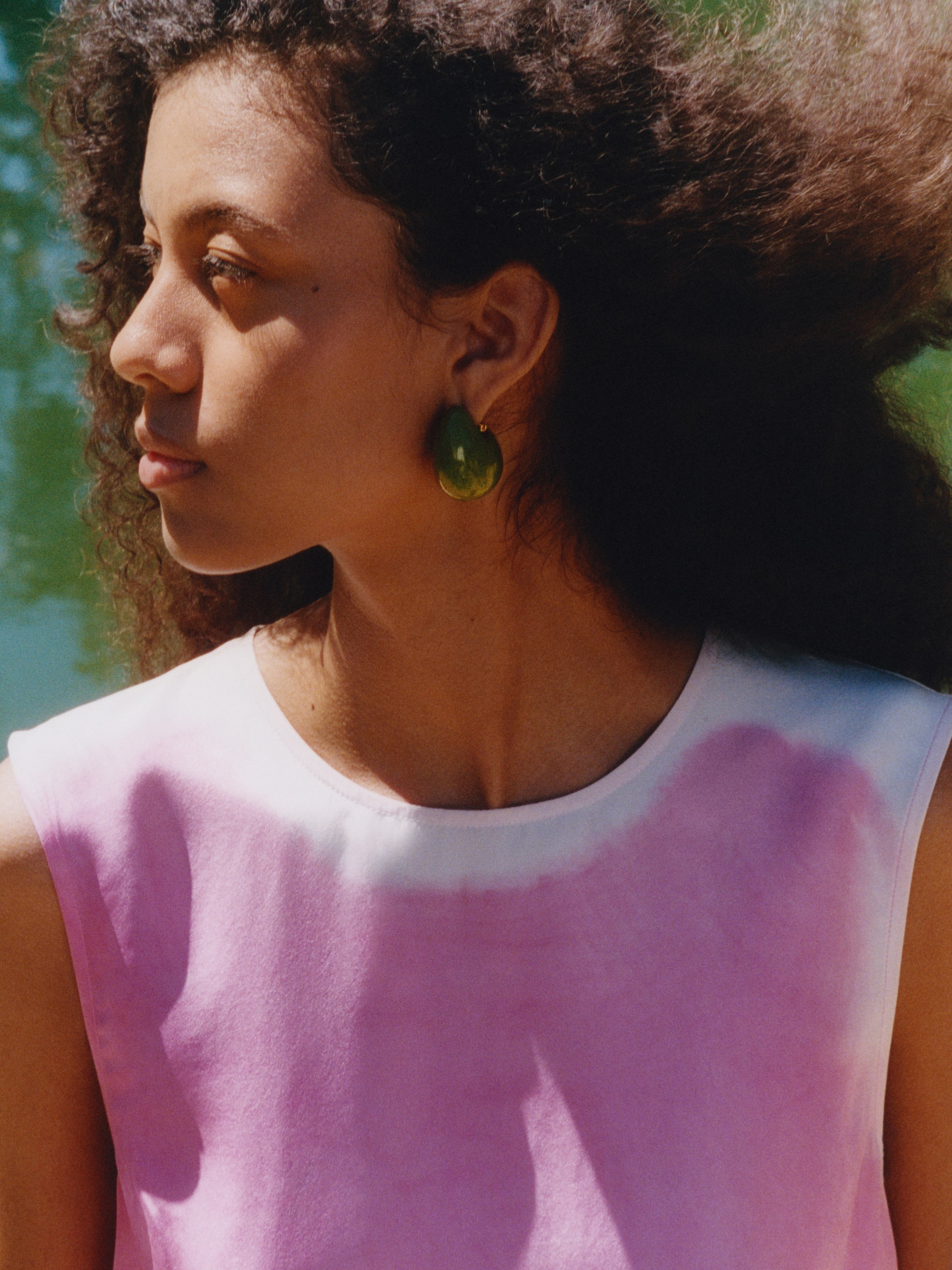 A woman with curly hair wearing green earrings and a pink and white top gazes to the side, with a blurred natural background.