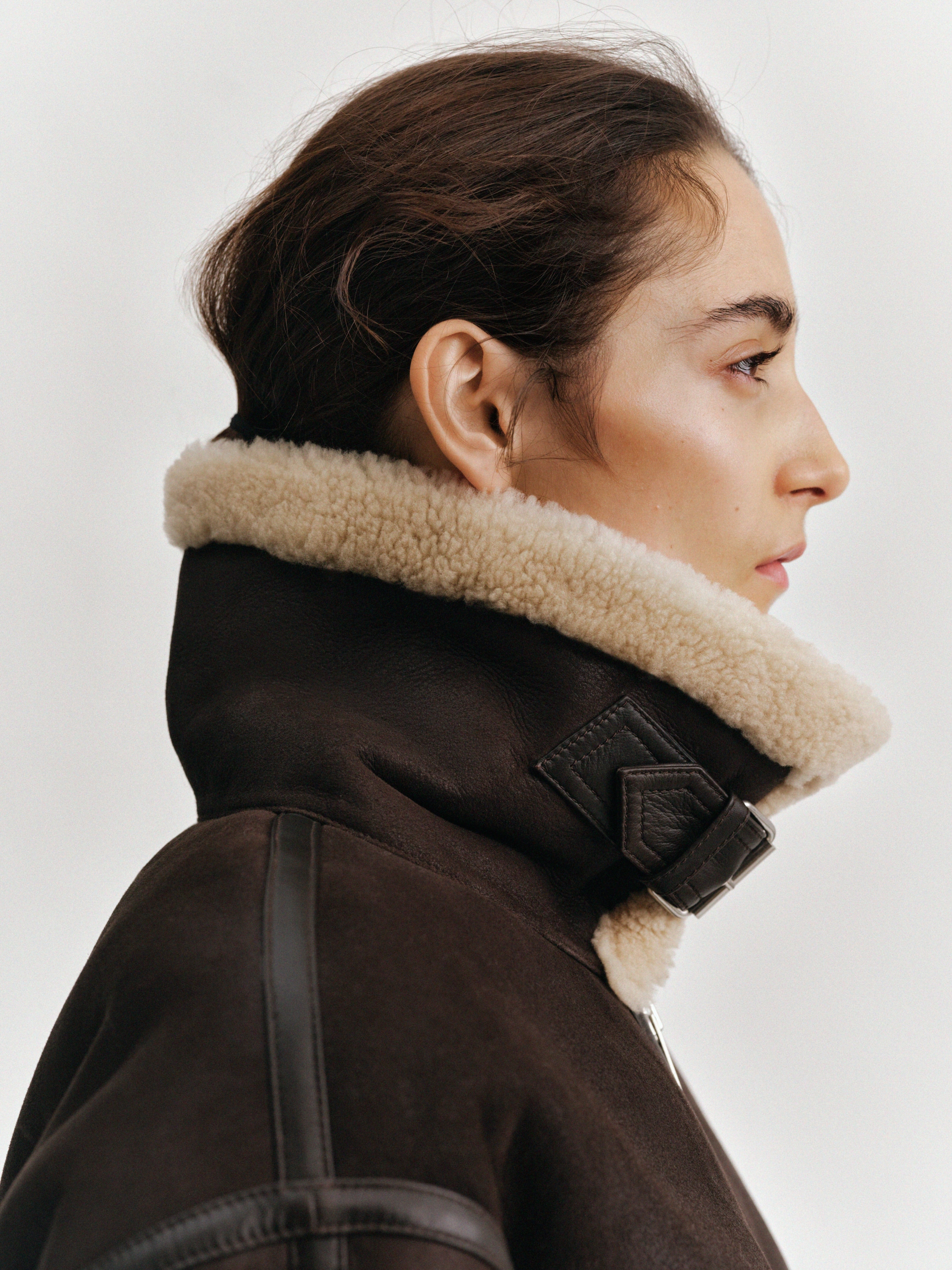 Profile of a person wearing a brown shearling jacket with a high collar, against a plain white background.