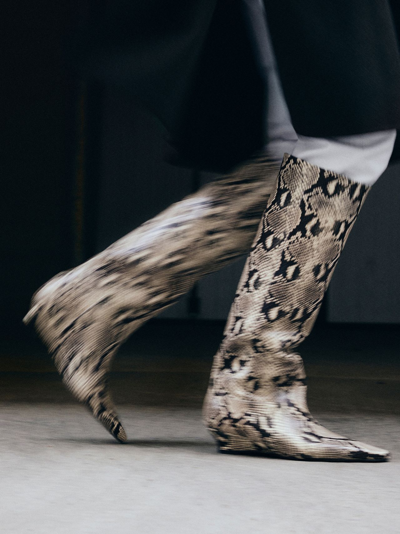 Person walking in knee-high, pointed-toe snakeskin boots, wearing a dark coat. The background is blurred, emphasizing motion.