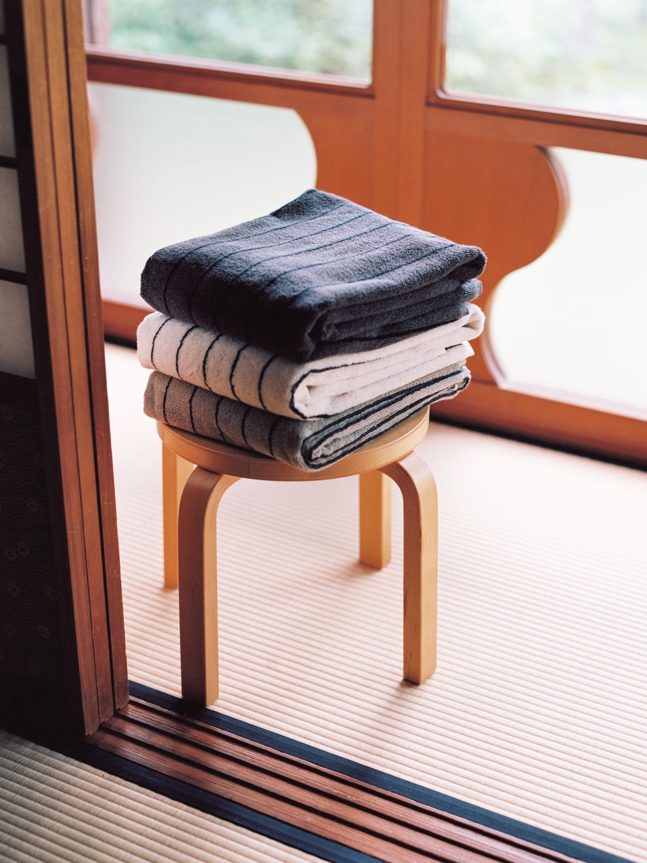A stack of folded towels in varying shades of gray sits on a wooden stool next to a sliding door in a sunlit room with tatami flooring.