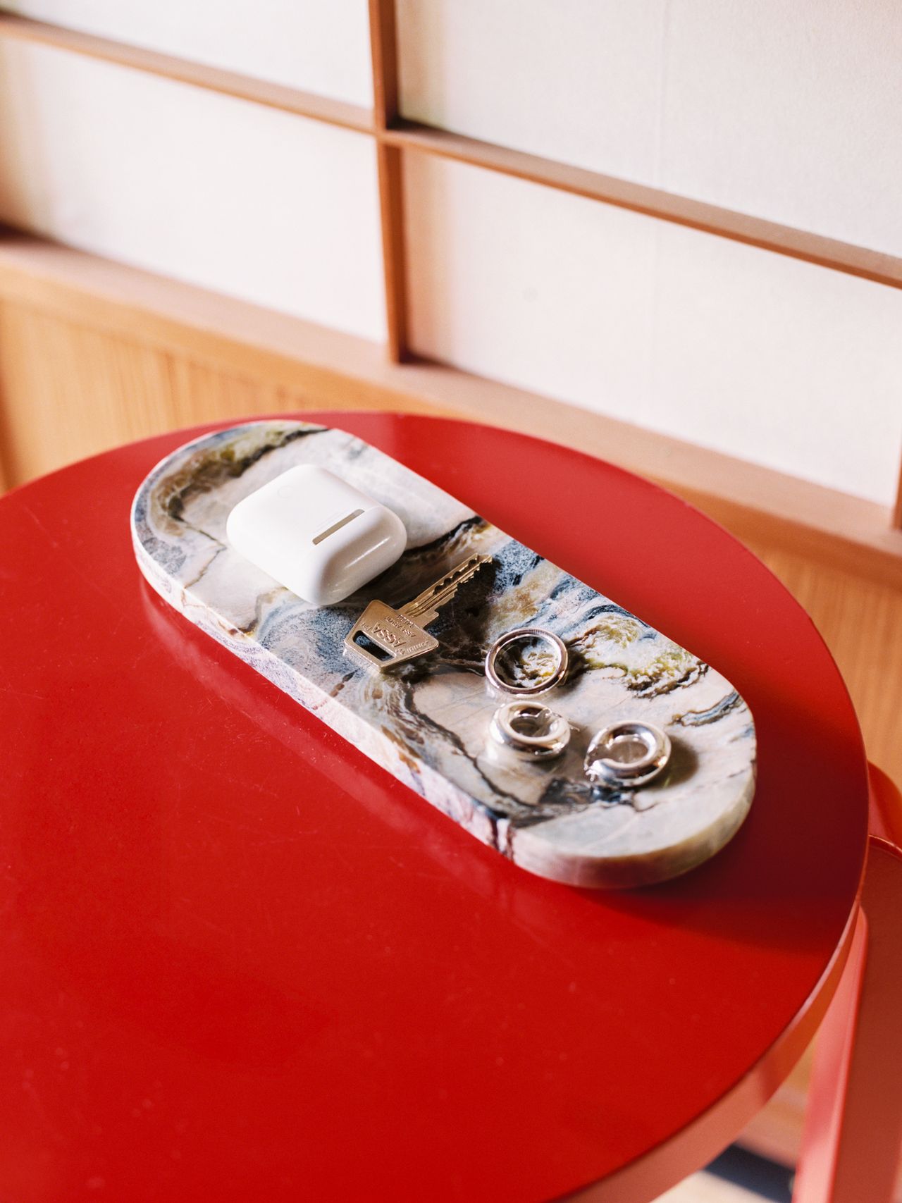 A marble tray with AirPods, a key, and rings sits on a round red table against a wooden and paper screen backdrop.