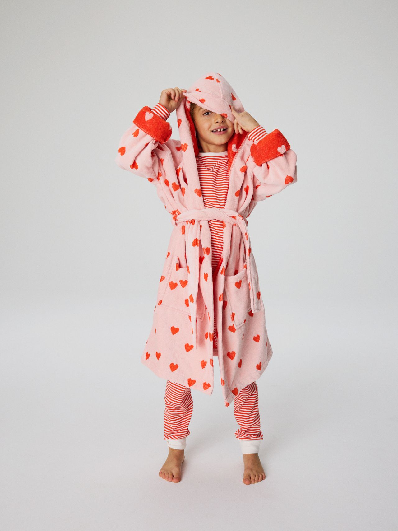 Child in heart-patterned robe and striped pajamas, playfully holding the hood, standing barefoot against a plain background.