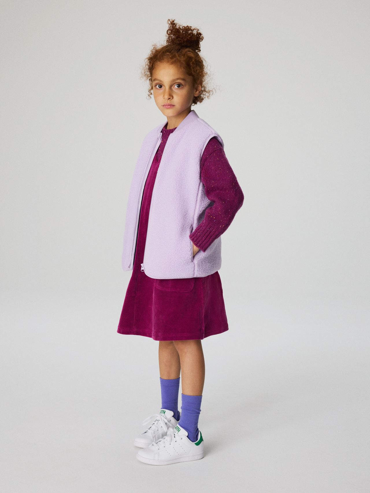 Young girl with curly hair wearing a purple dress, lavender vest, and sneakers, standing against a plain background.