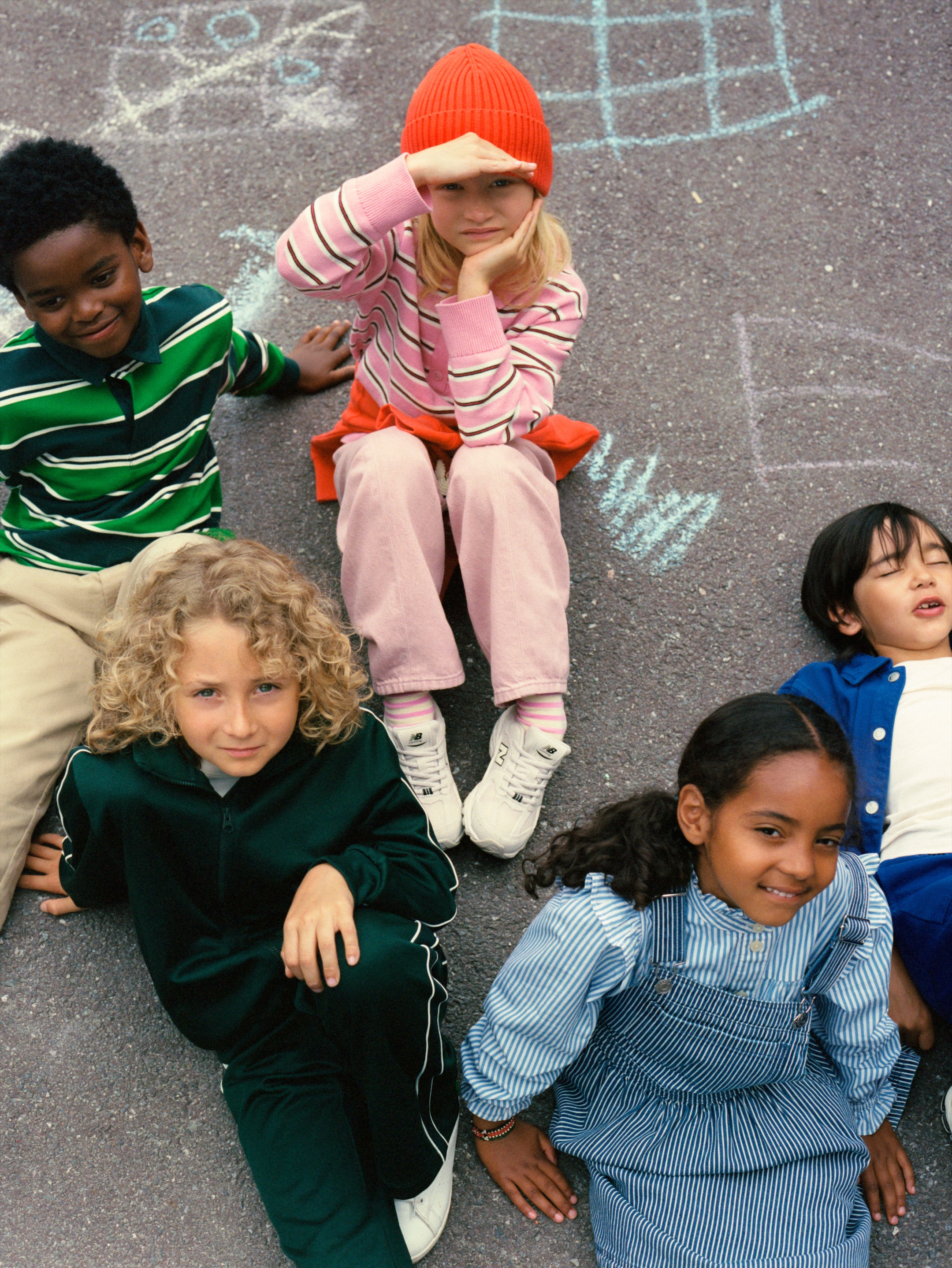 Five children sitting on pavement with colorful chalk drawings. One wears a red hat and pink clothes, others smile and relax in playful poses.