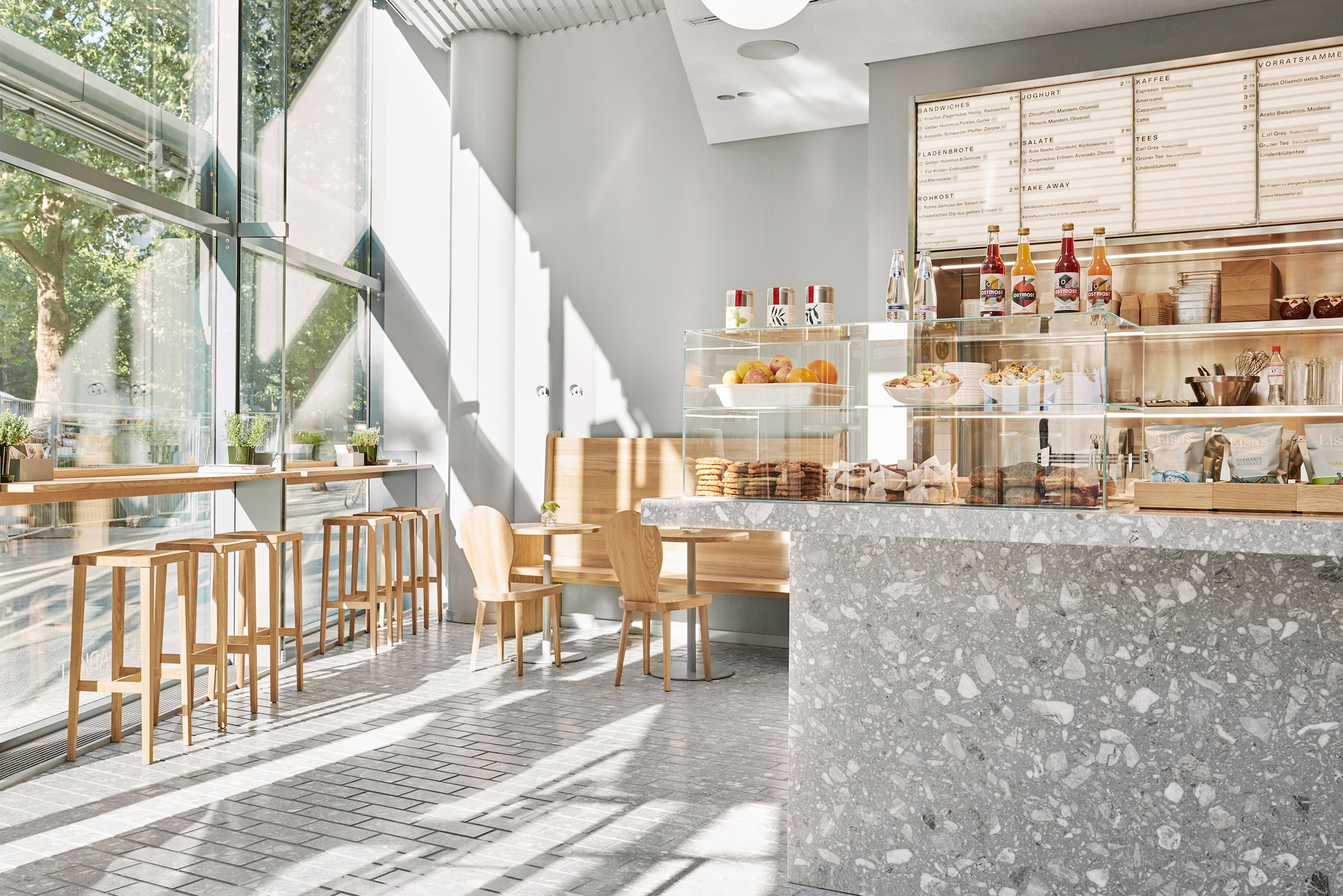 Sunlit cafe interior with wooden furniture, large glass windows, and a counter displaying pastries and drinks.