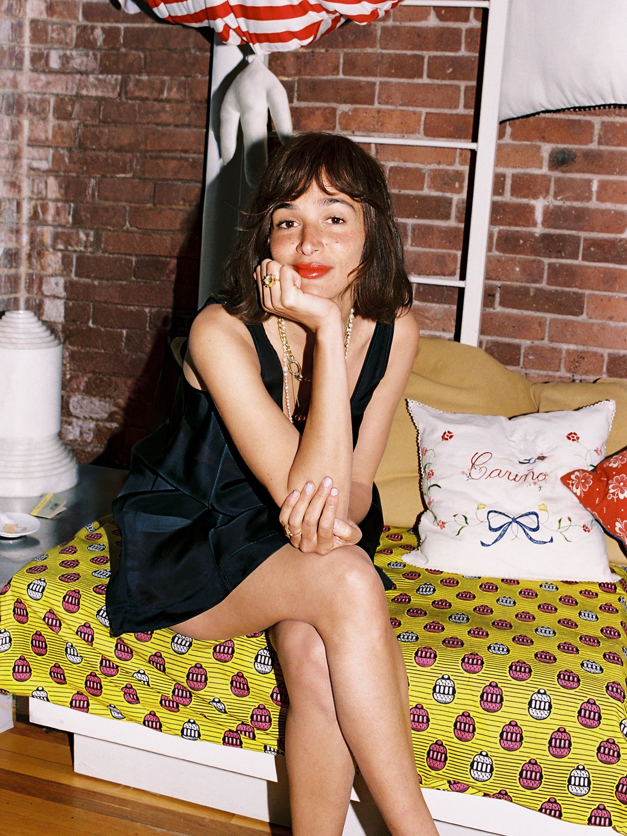 A woman in a black dress sits on a bed with colorful bedding, in a room with exposed brick walls.
