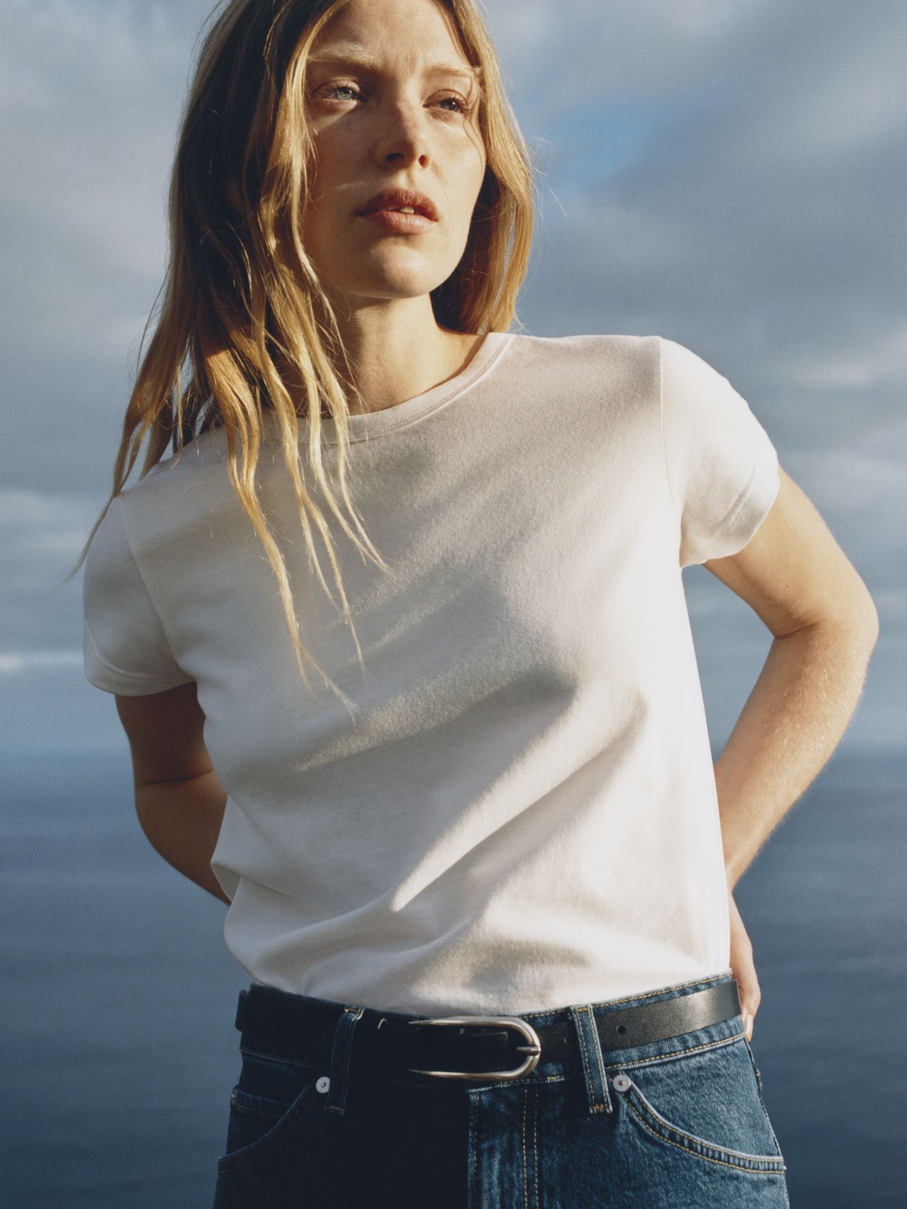 Person with long hair in a white t-shirt and jeans stands by the sea, looking thoughtful under a cloudy sky.
