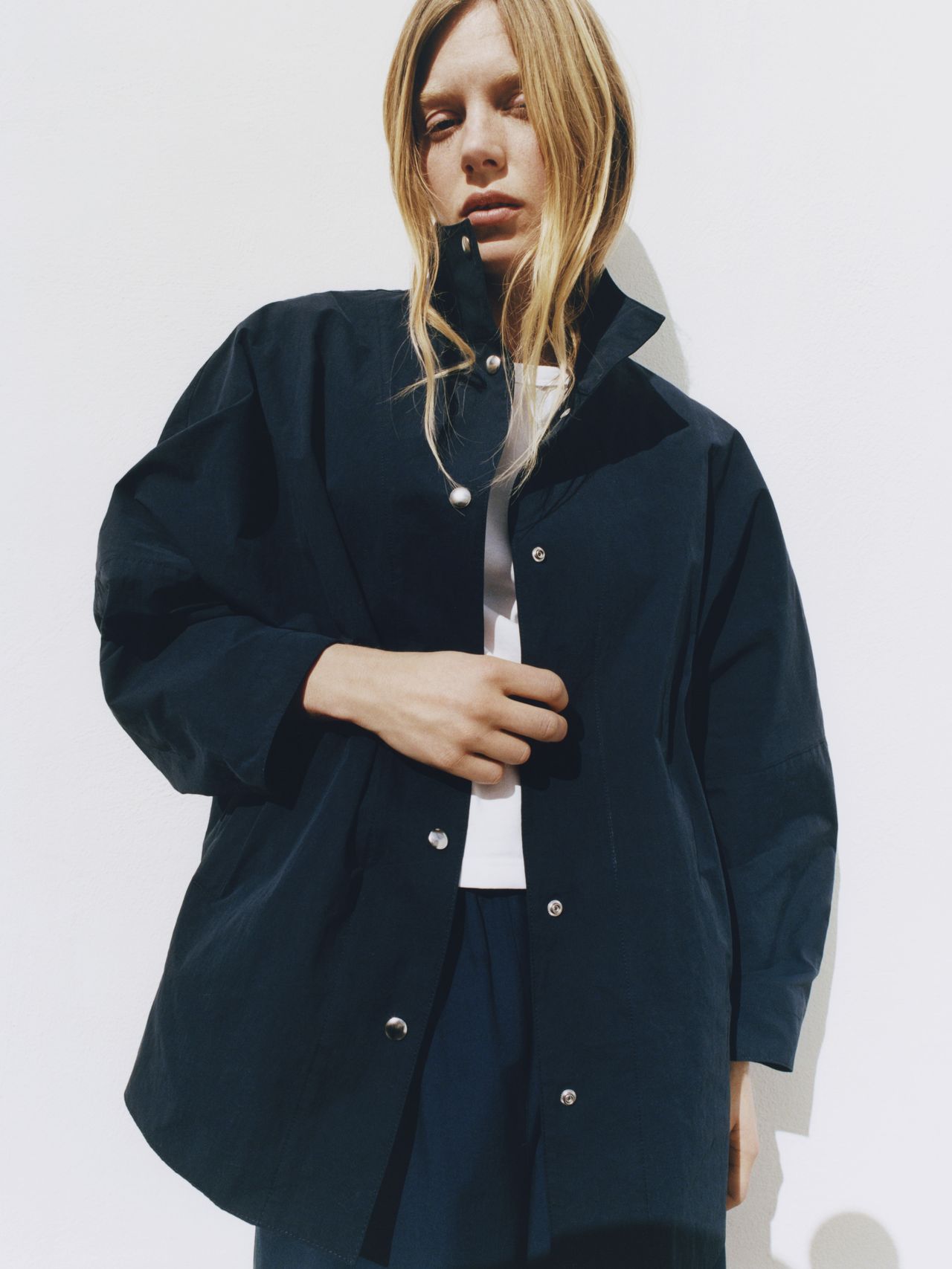 Person in a navy jacket with silver buttons stands against a white background, with long blonde hair partially covering the face.