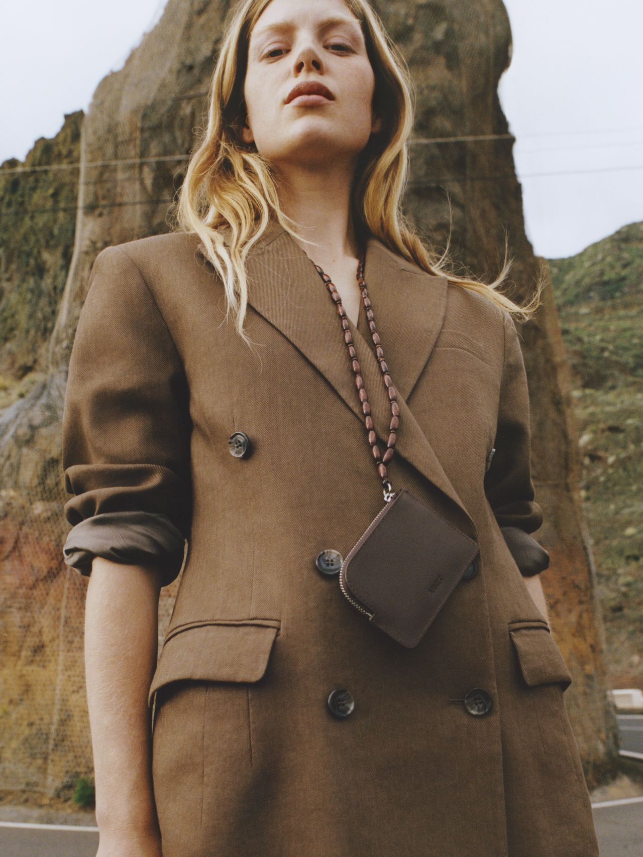 Person in a brown double-breasted blazer stands confidently outdoors with rocky cliffs and greenery in the background.