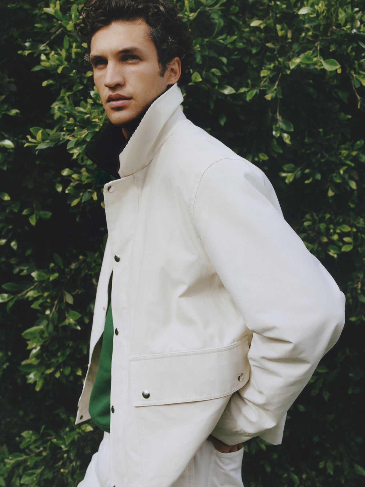 Person in a white jacket with a green shirt stands against a background of lush green leaves, looking to the side.